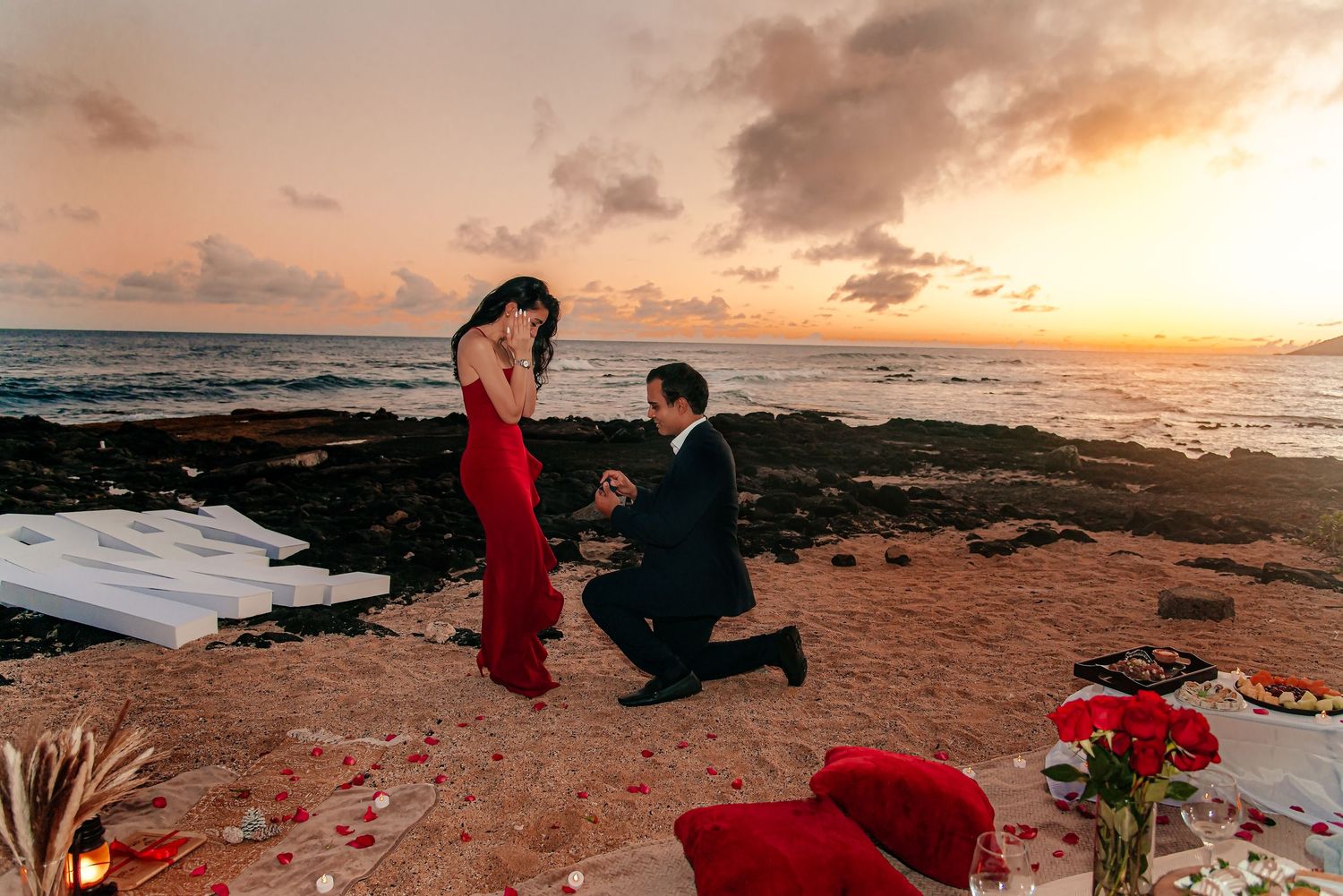 Man proposing to woman in red dress on beach at sunset with roses and romantic decorations.