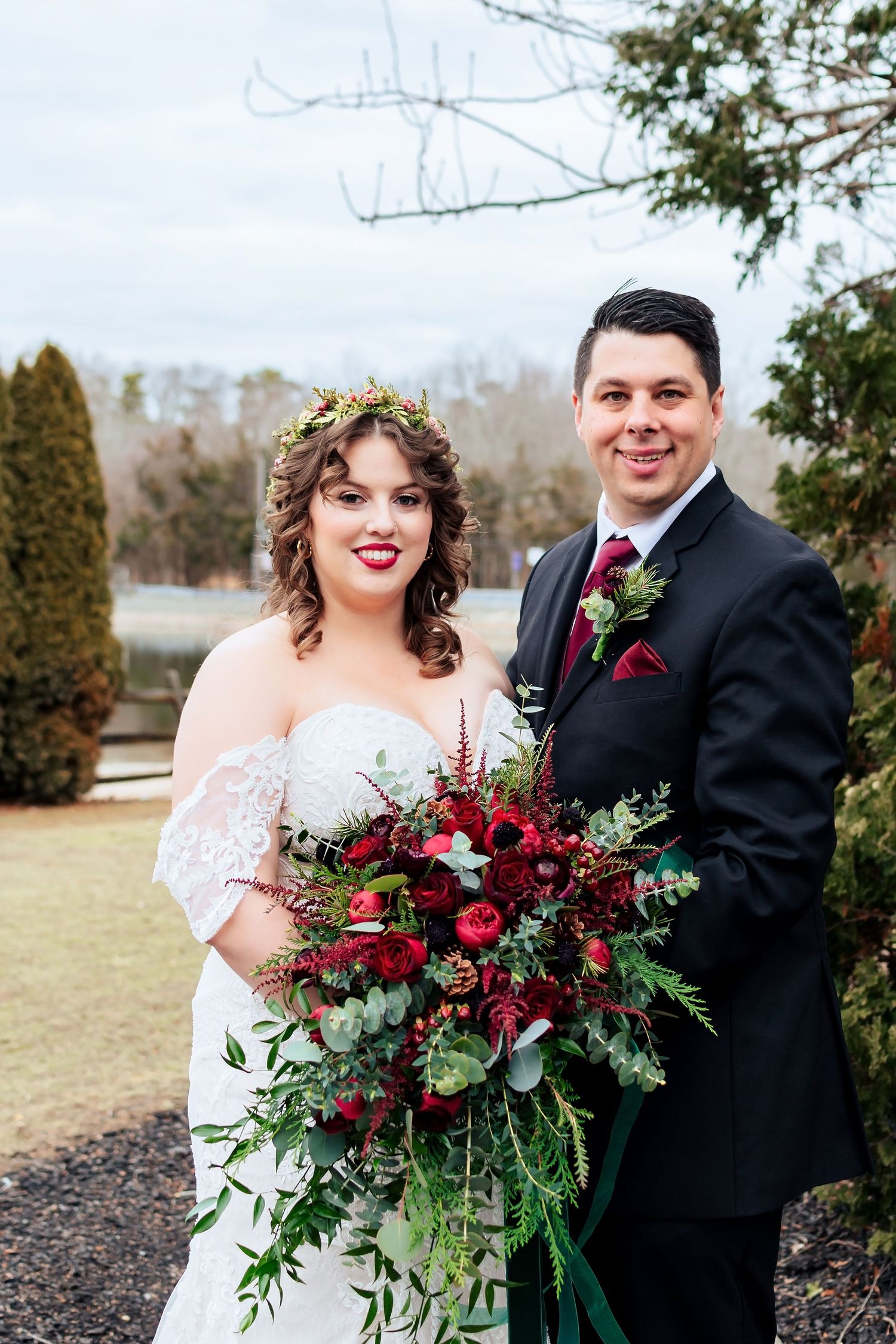 A bride wearing an off-shoulder dress and floral crown holds a lush burgundy and greenery bouquet next to a groom in black suit.