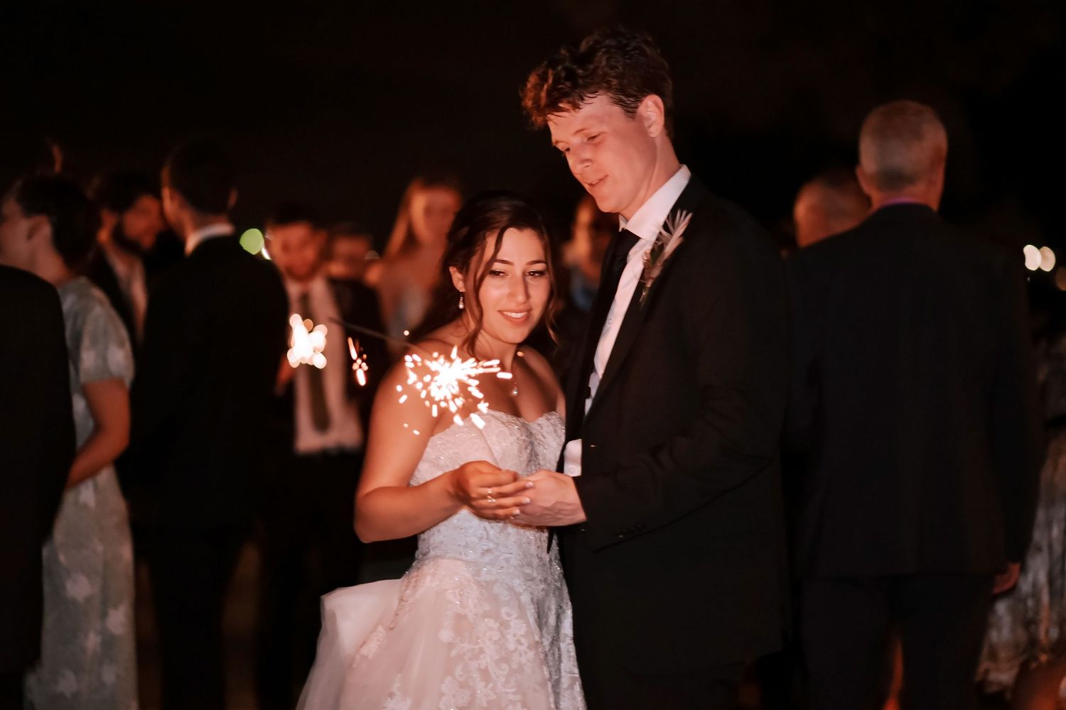 Bride and groom holding sparklers together at wedding reception with guests in background at night.