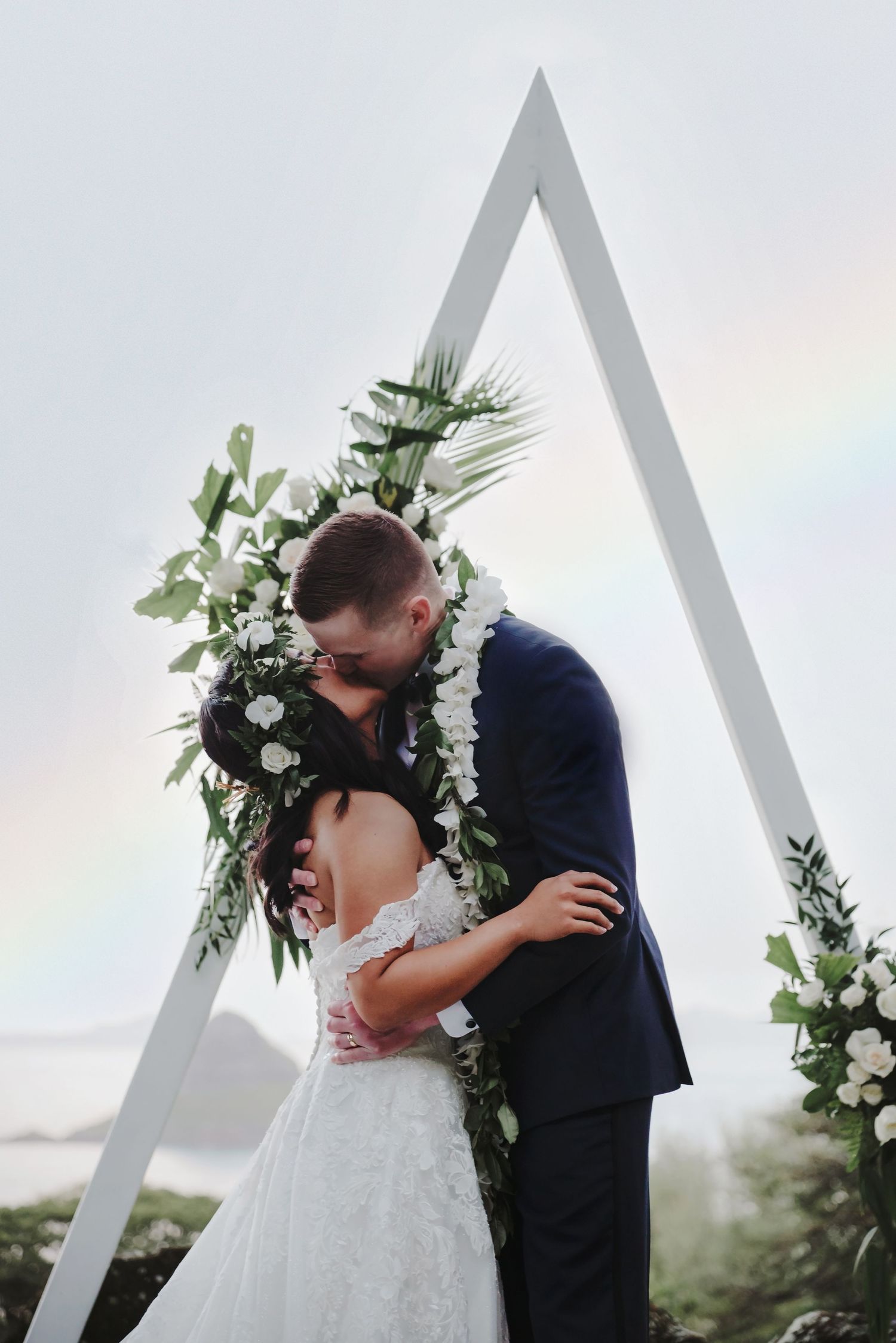A romantic wedding kiss sequence between newlyweds under a white arch decorated with lush greenery and white flowers.
