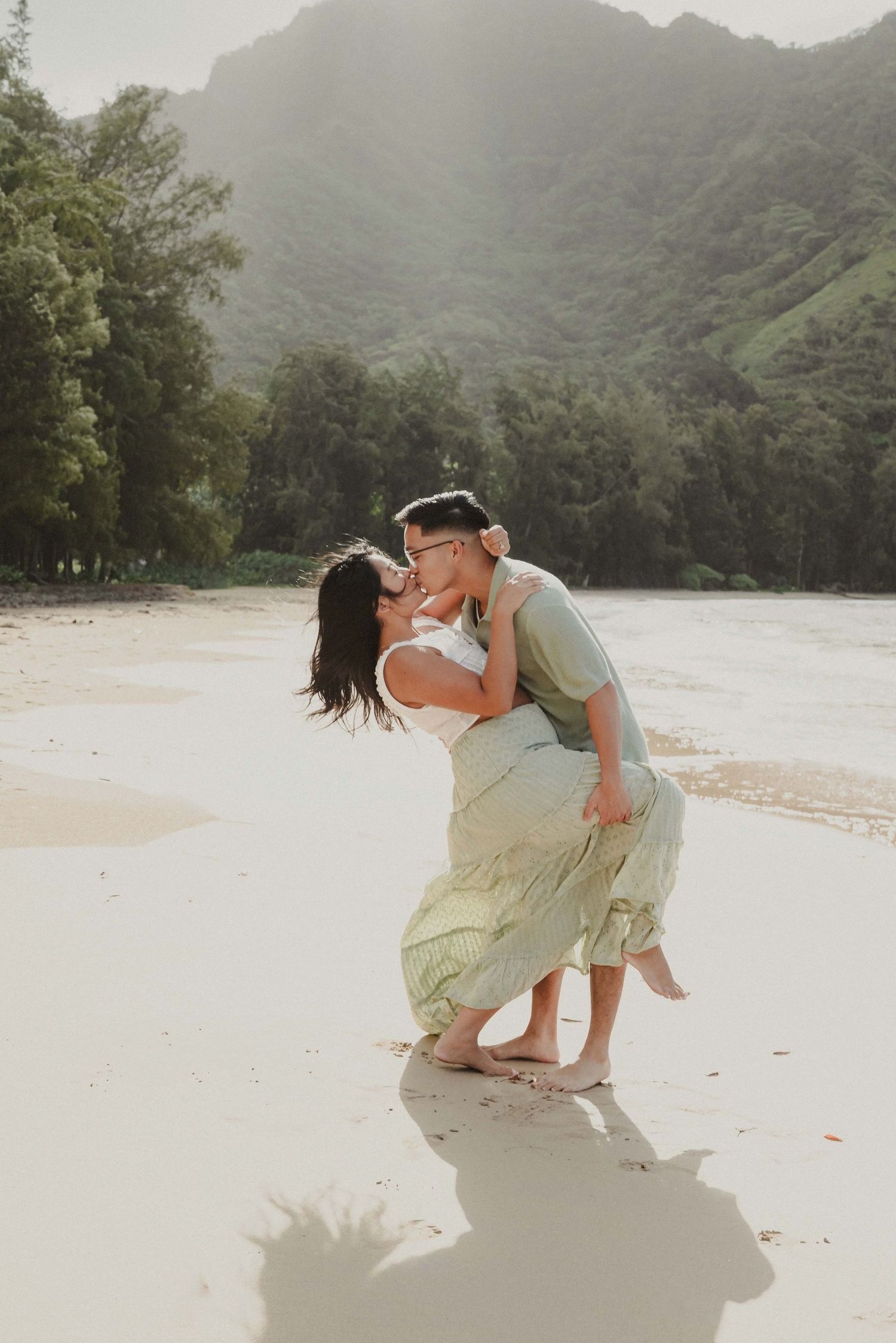 A romantic couple embraces and dances together on a misty beach with lush mountains in the background.