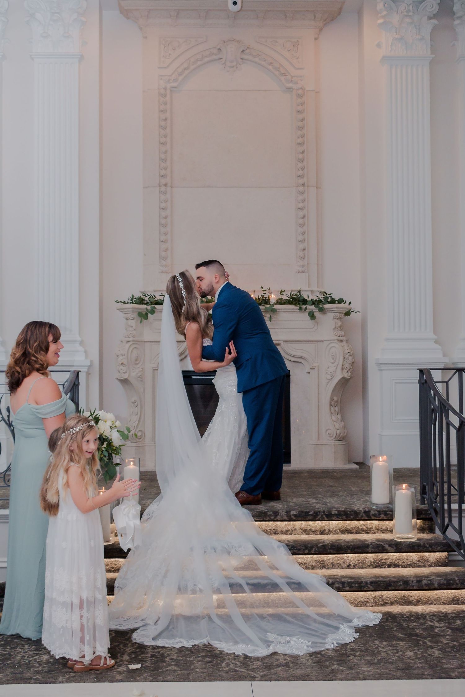 Bride and groom share first kiss at elegant white fireplace altar, with bridesmaid and flower girl nearby.