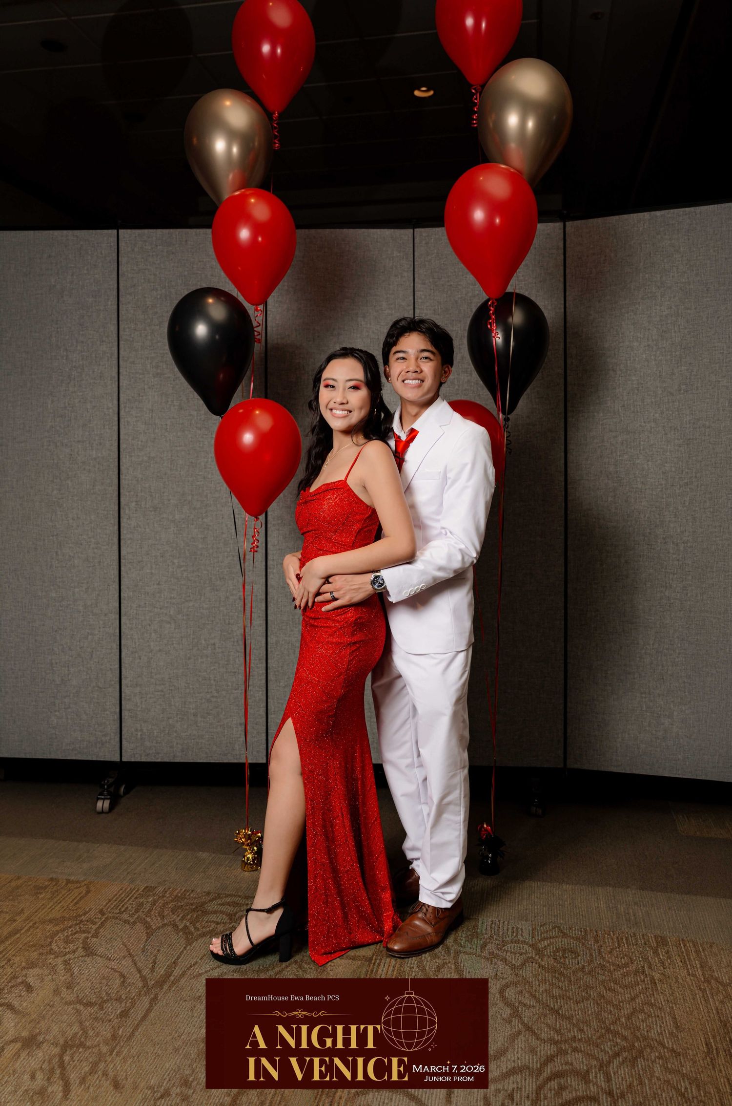Couple in red dress and white suit posing with red, black, and gold balloons at A Night in Venice event.
