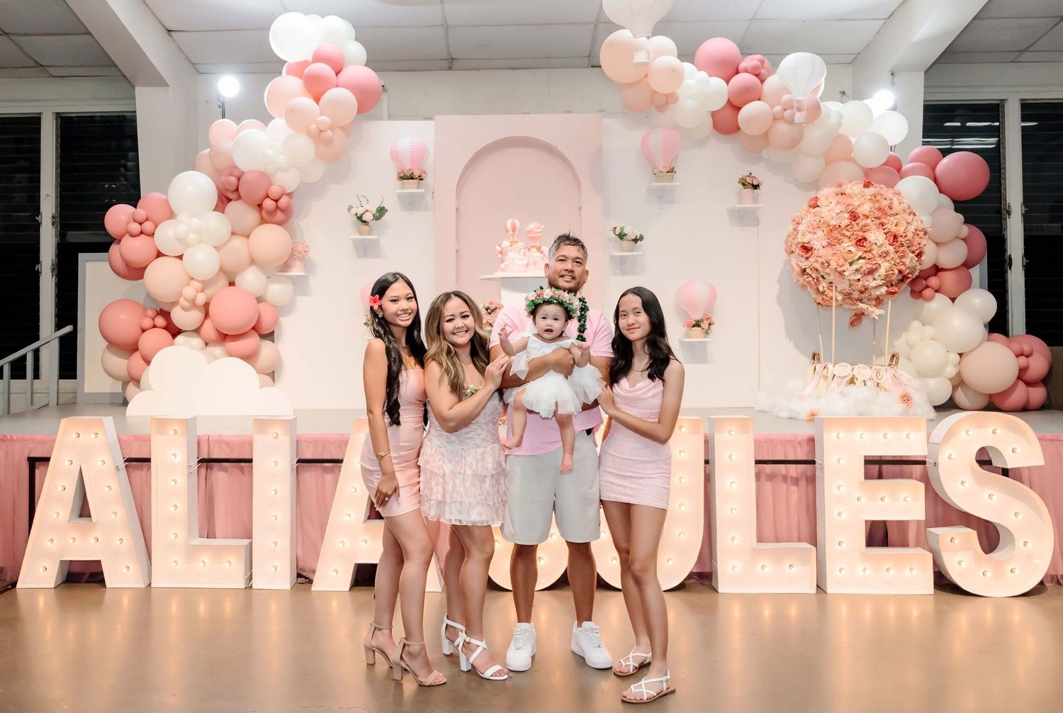 Family posing in front of pink balloon arch and large letter decorations at a birthday party celebration.