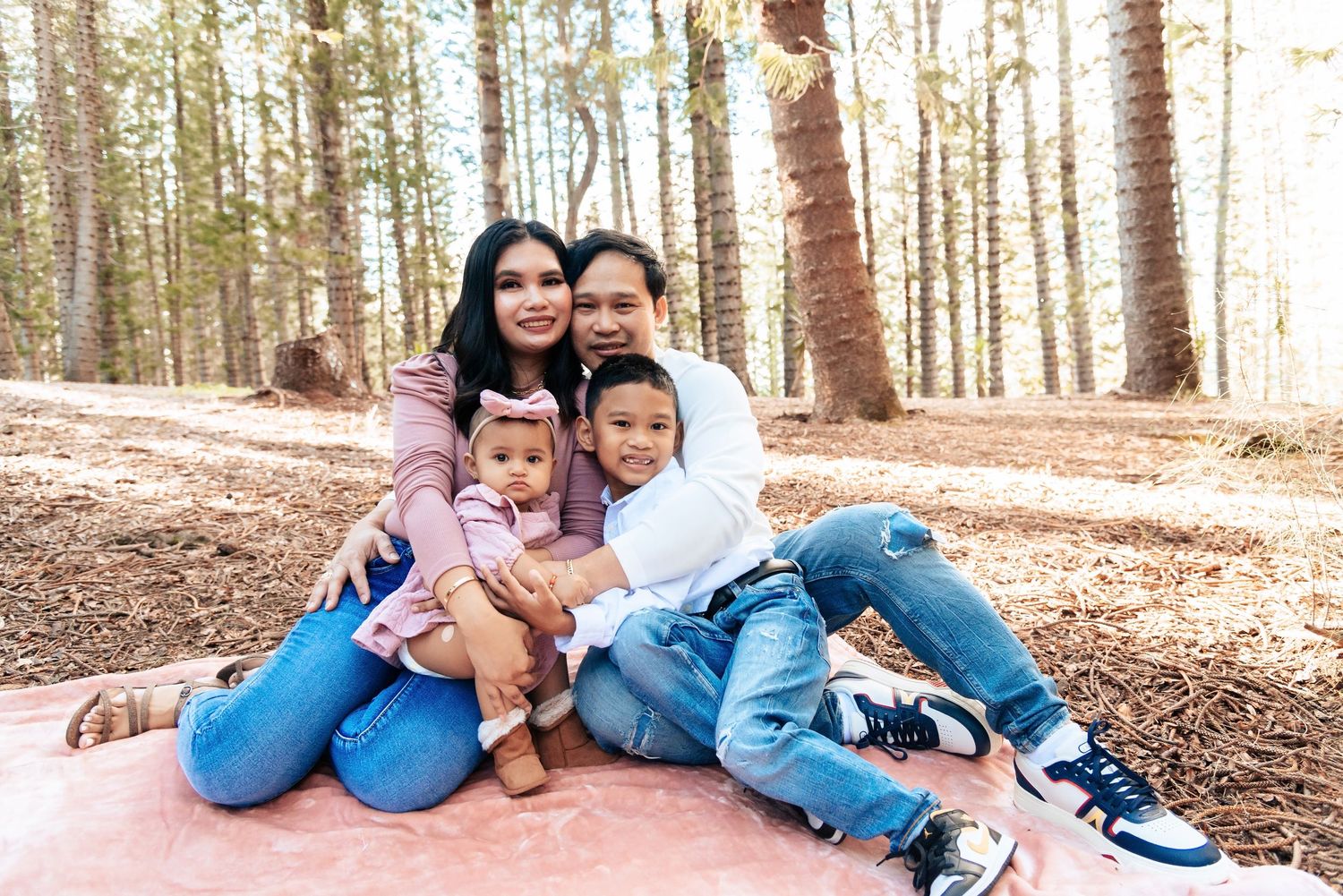 Family of four posing together on a blanket in a sunlit forest, smiling warmly for a portrait photo.