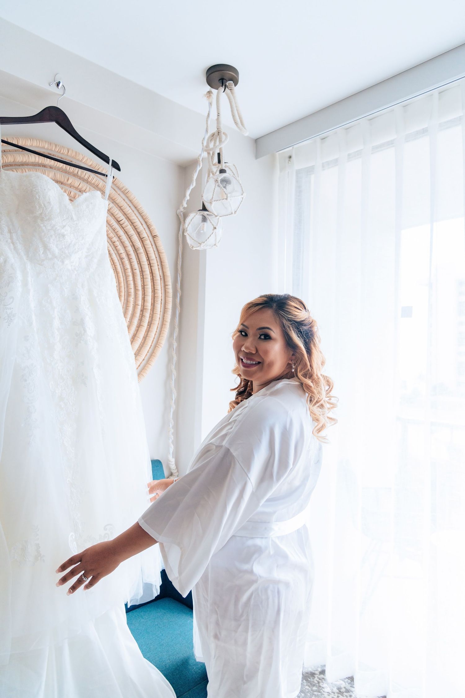 Bridal preparation scene with white wedding dress hanging by window as someone in white robe stands nearby.