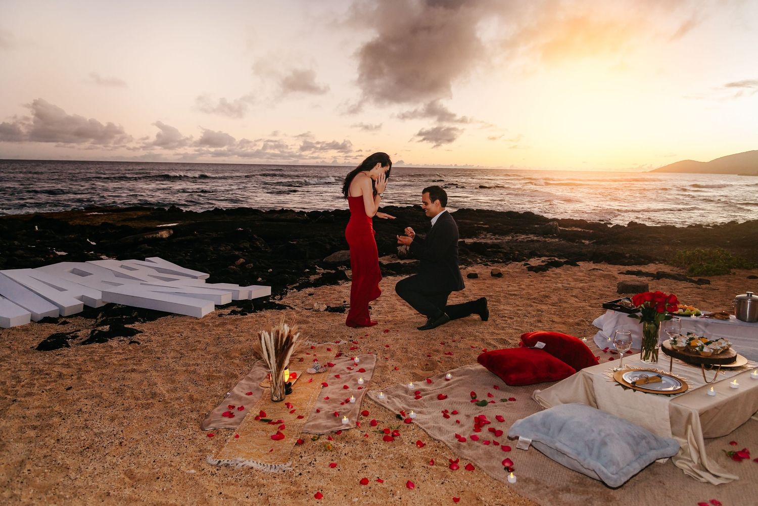 Man proposes to woman on sandy beach at sunset with rose petals and romantic dinner setup in Hawaii.