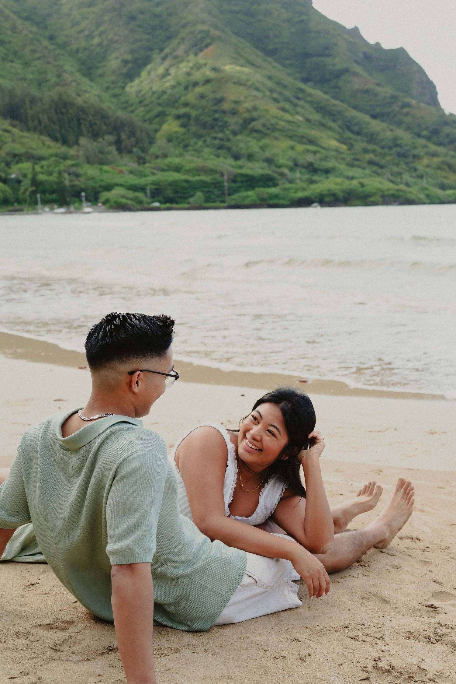 Couple relaxing on sandy beach with lush green mountains and calm ocean water in the background.