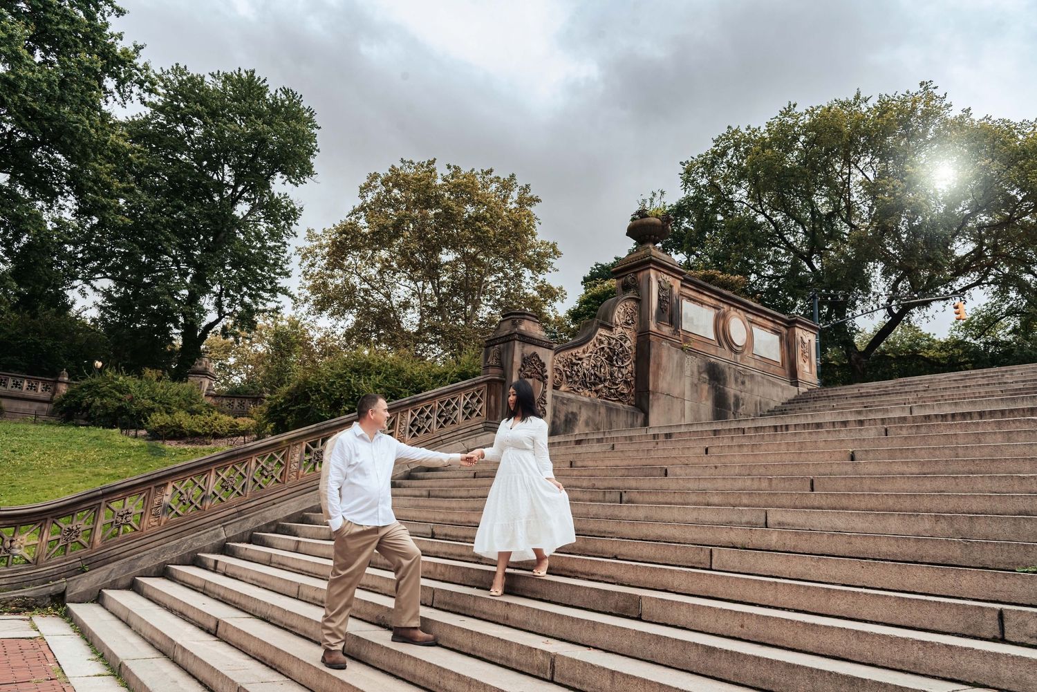 A couple in white outfits walks up stone amphitheater steps on an overcast day in a park setting.
