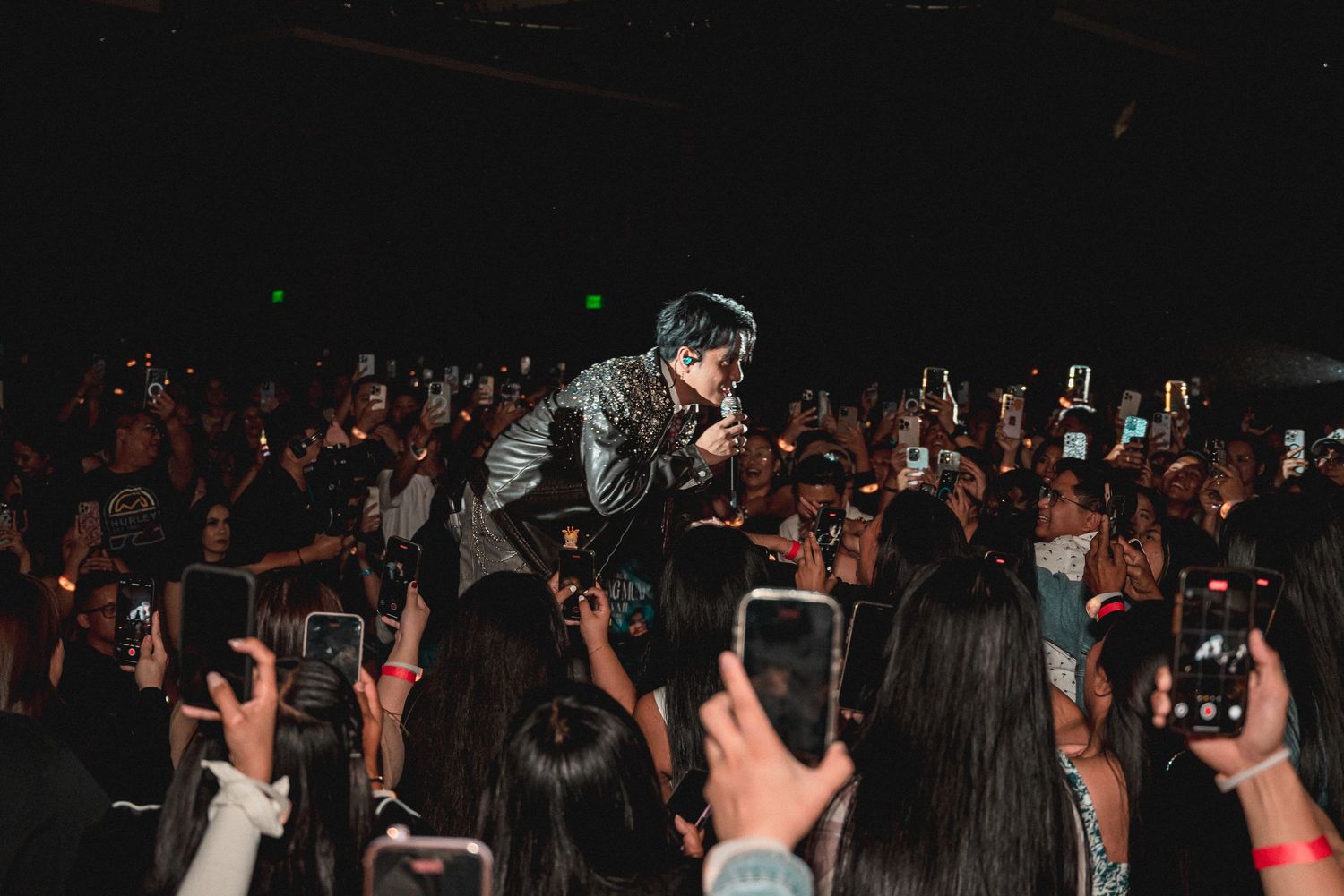 A performer sings on stage surrounded by an enthusiastic crowd holding up smartphones at a dark concert venue.