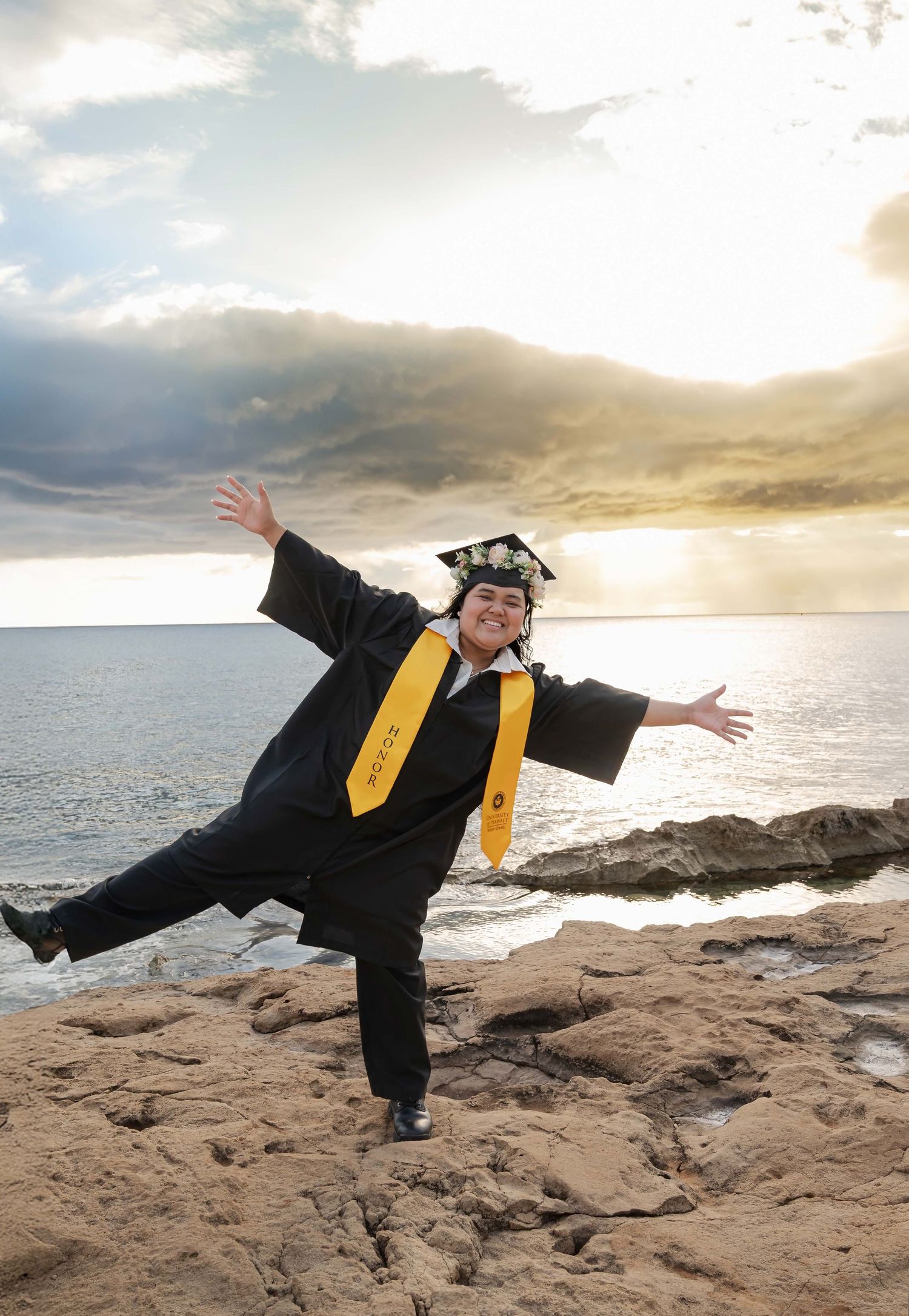 Graduate in cap and gown poses joyfully on rocky shoreline at sunset with arms outstretched and one leg raised.