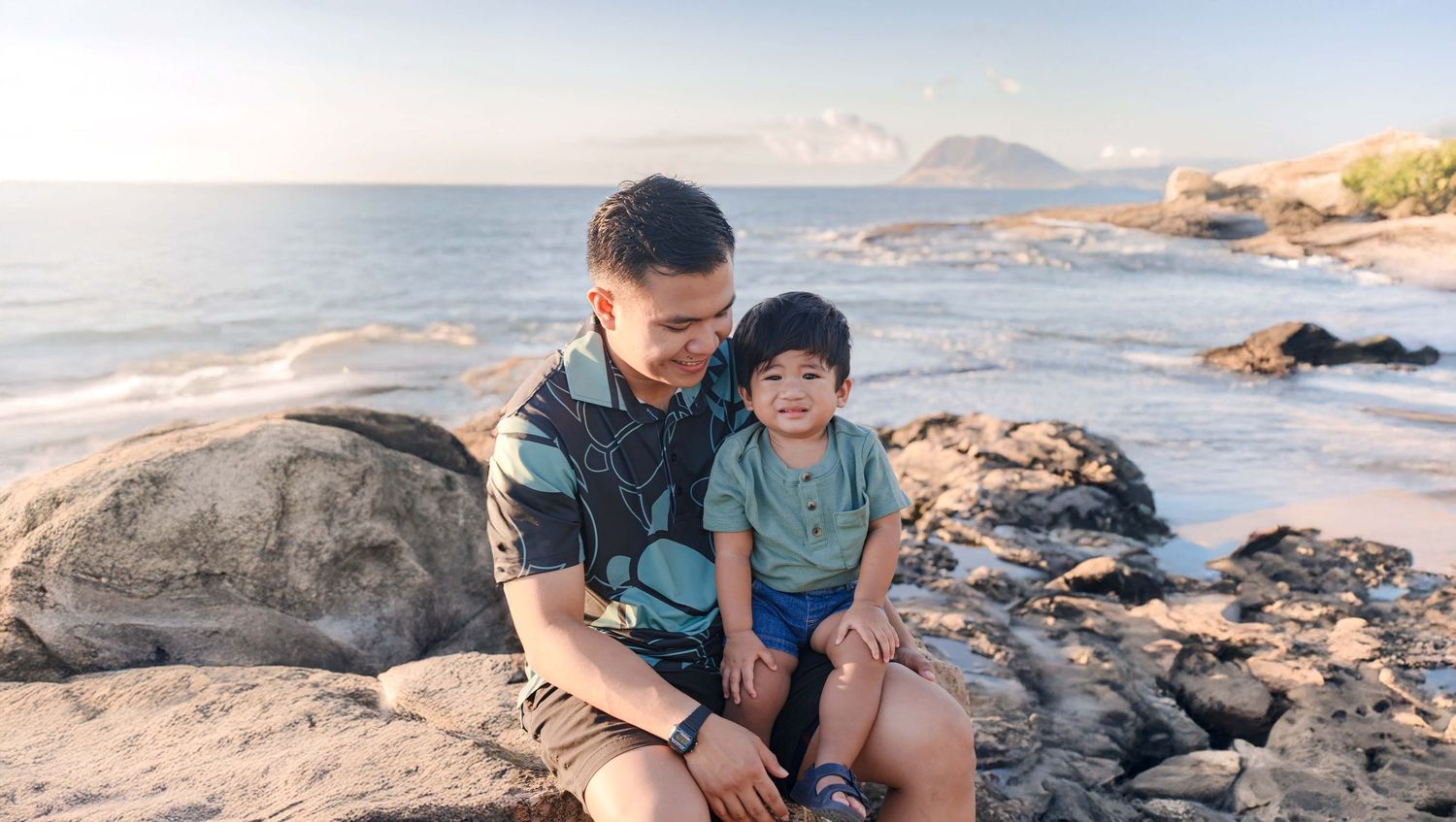 Father and young child sitting on rocky beach at sunset with ocean and mountain island in background.