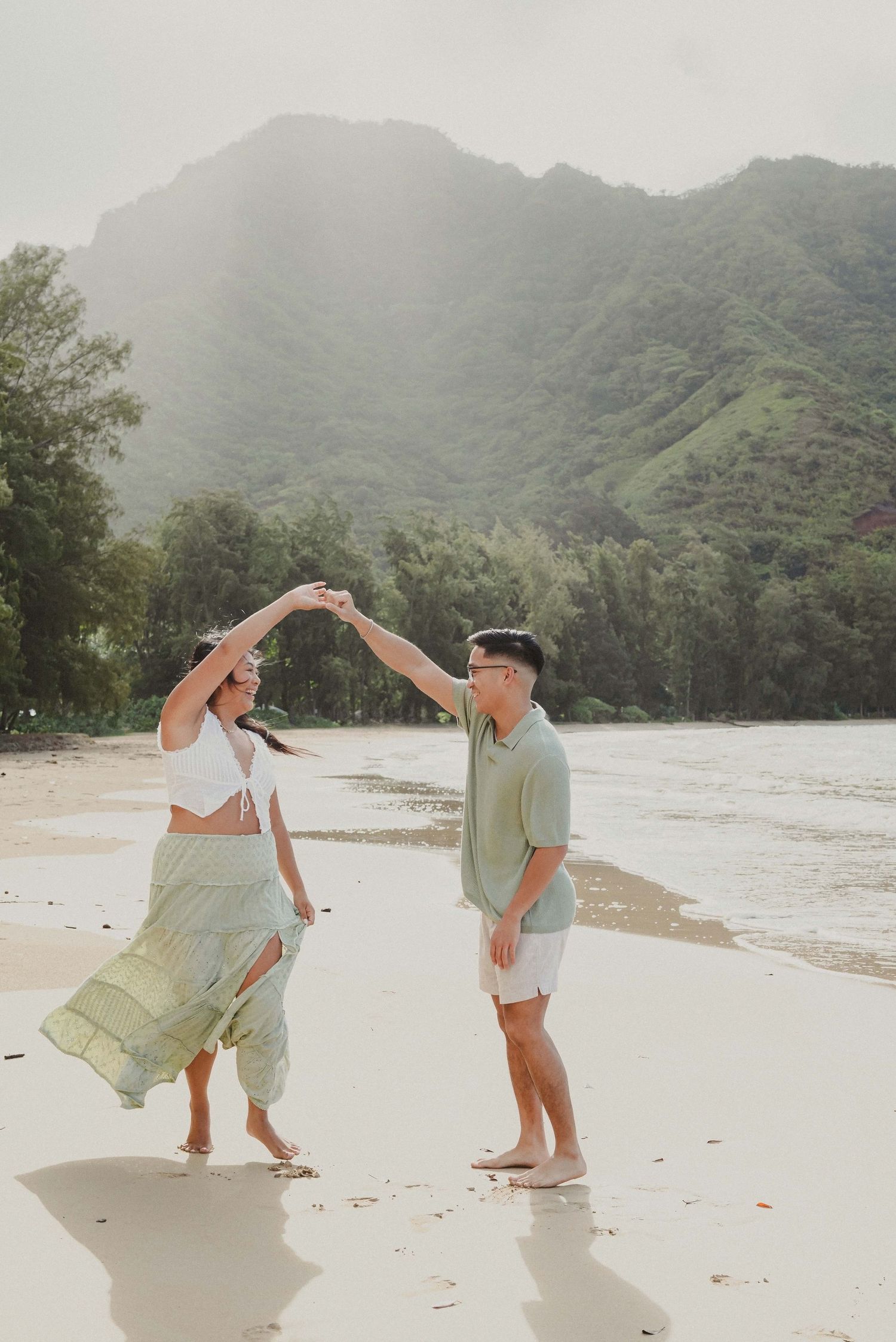 Couple dancing on a tropical beach with lush green mountains in the background on a misty day.