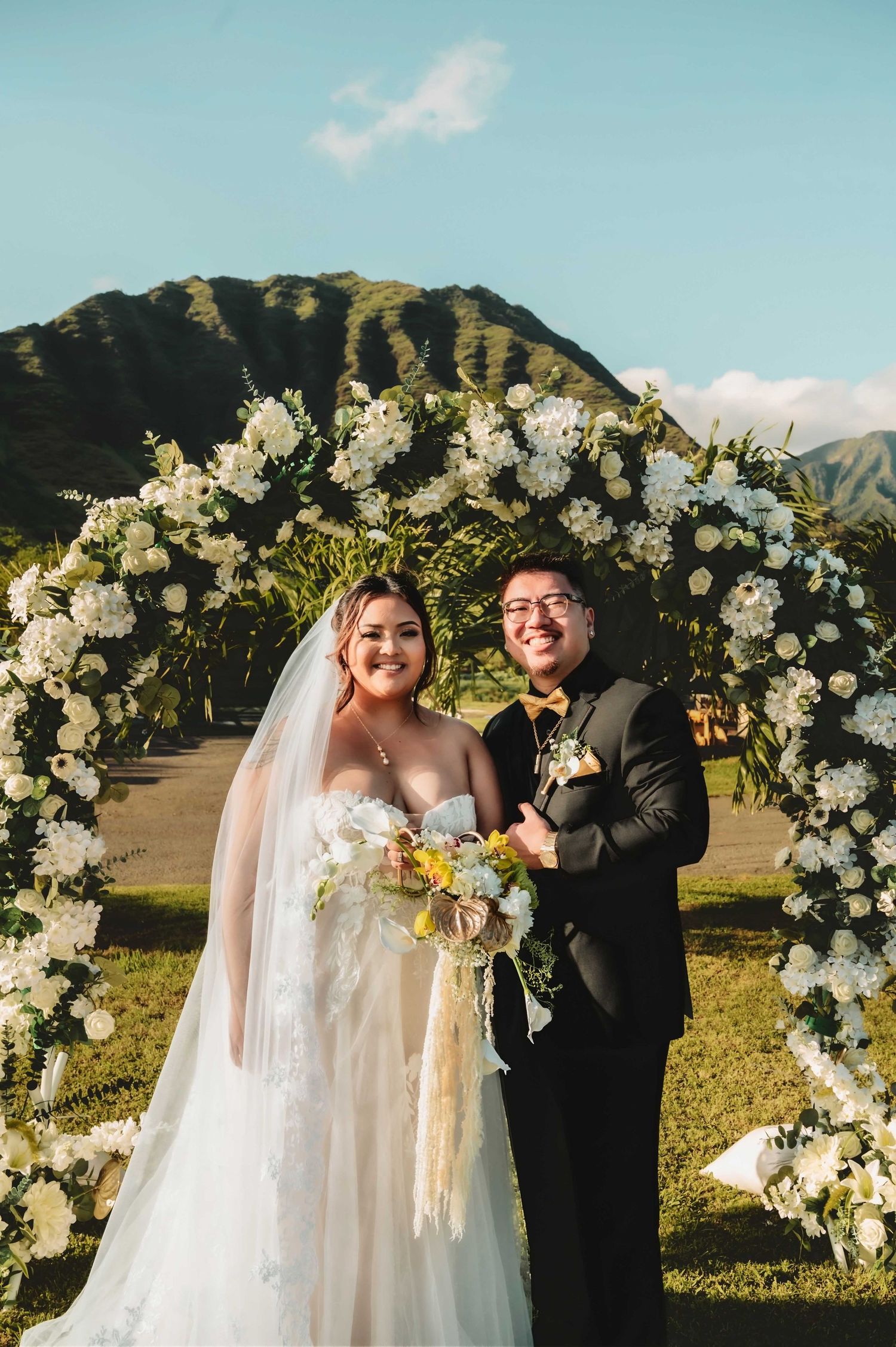 Bride and groom smiling under a white floral arch with mountain backdrop at an outdoor Hawaii wedding ceremony.