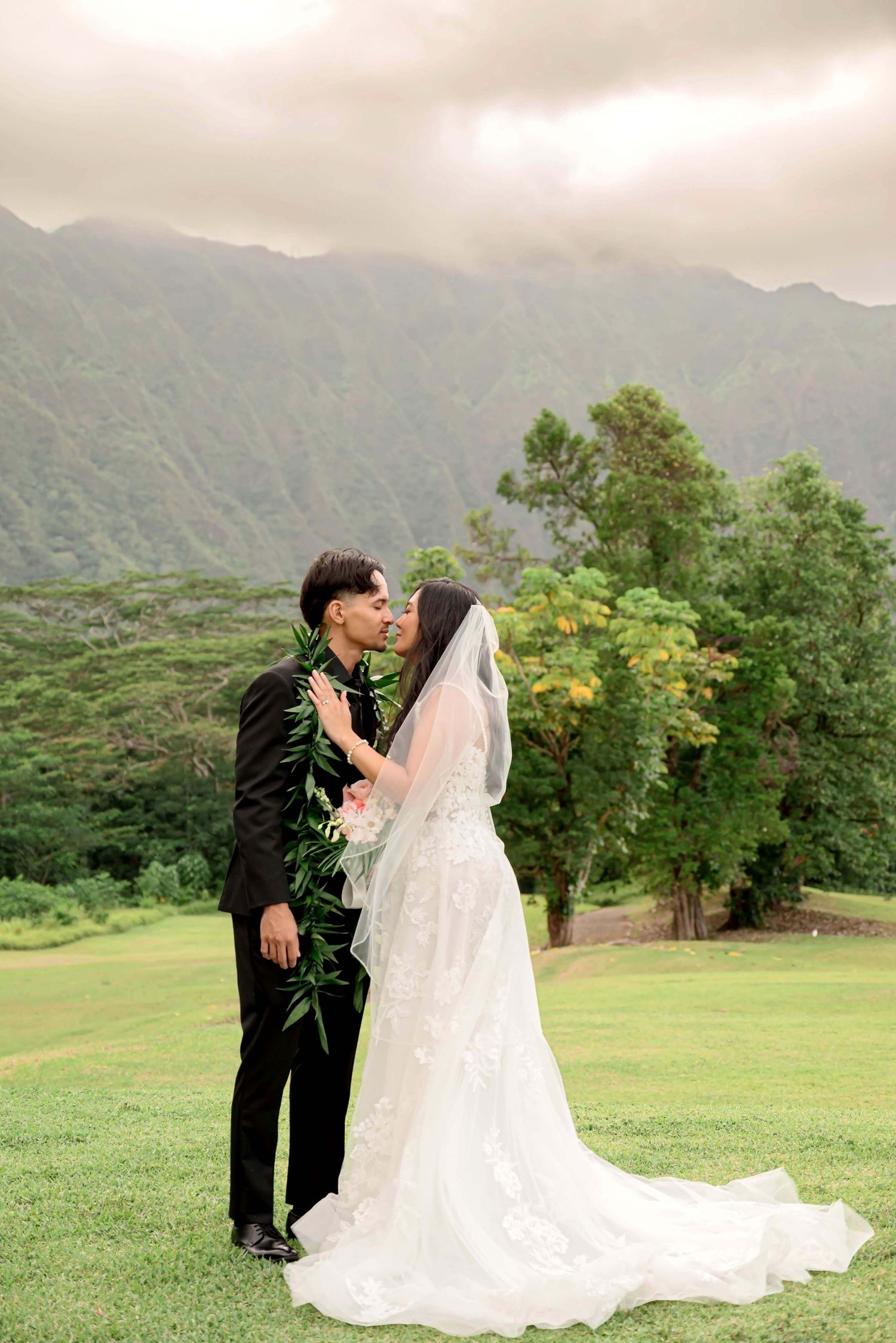 A romantic wedding couple shares a kiss on a lush green lawn with misty mountains creating a dramatic backdrop in Hawaii.