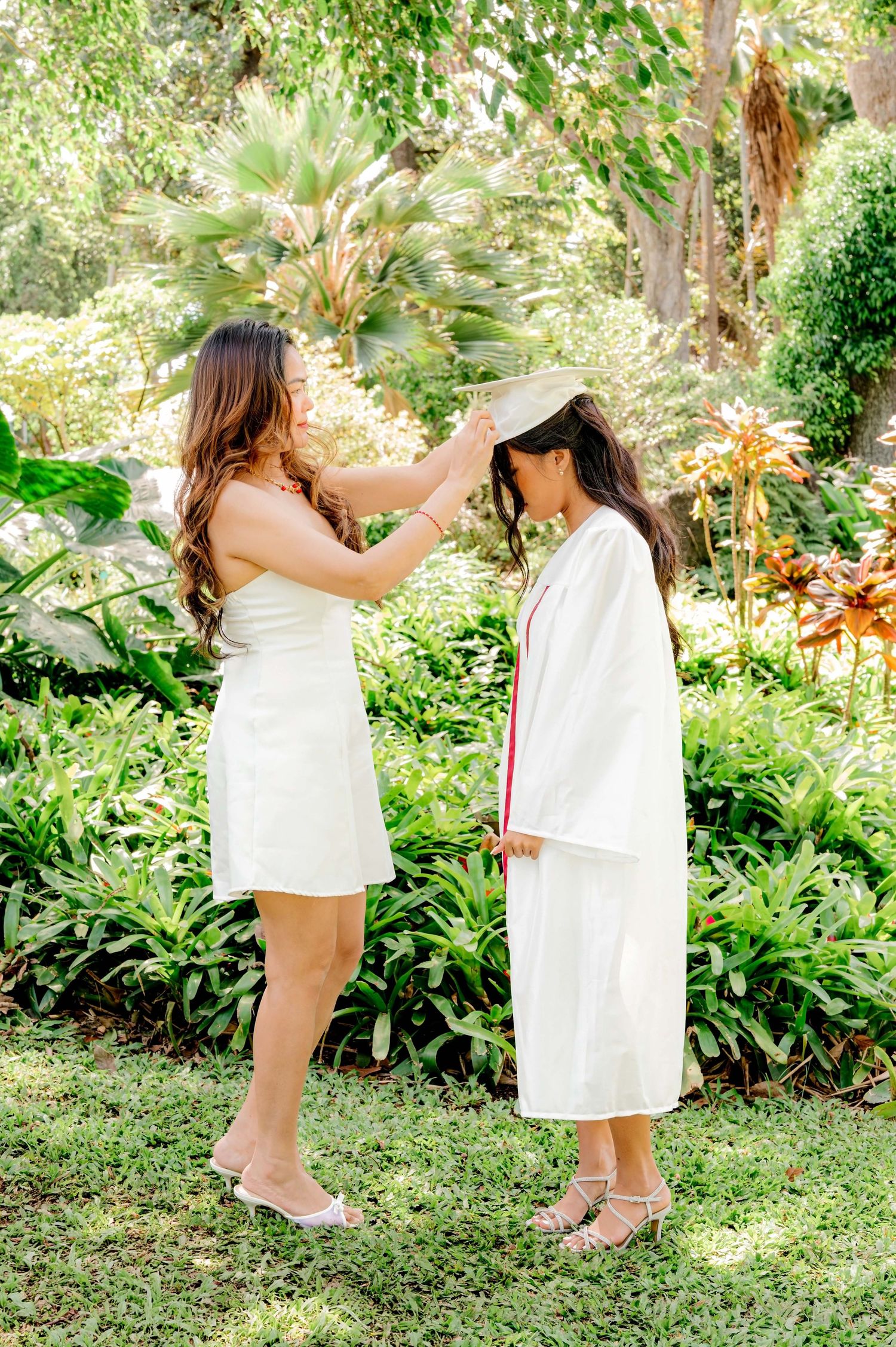 Woman in white dress adjusting graduation cap on graduate in white cap and gown outdoors in lush garden.