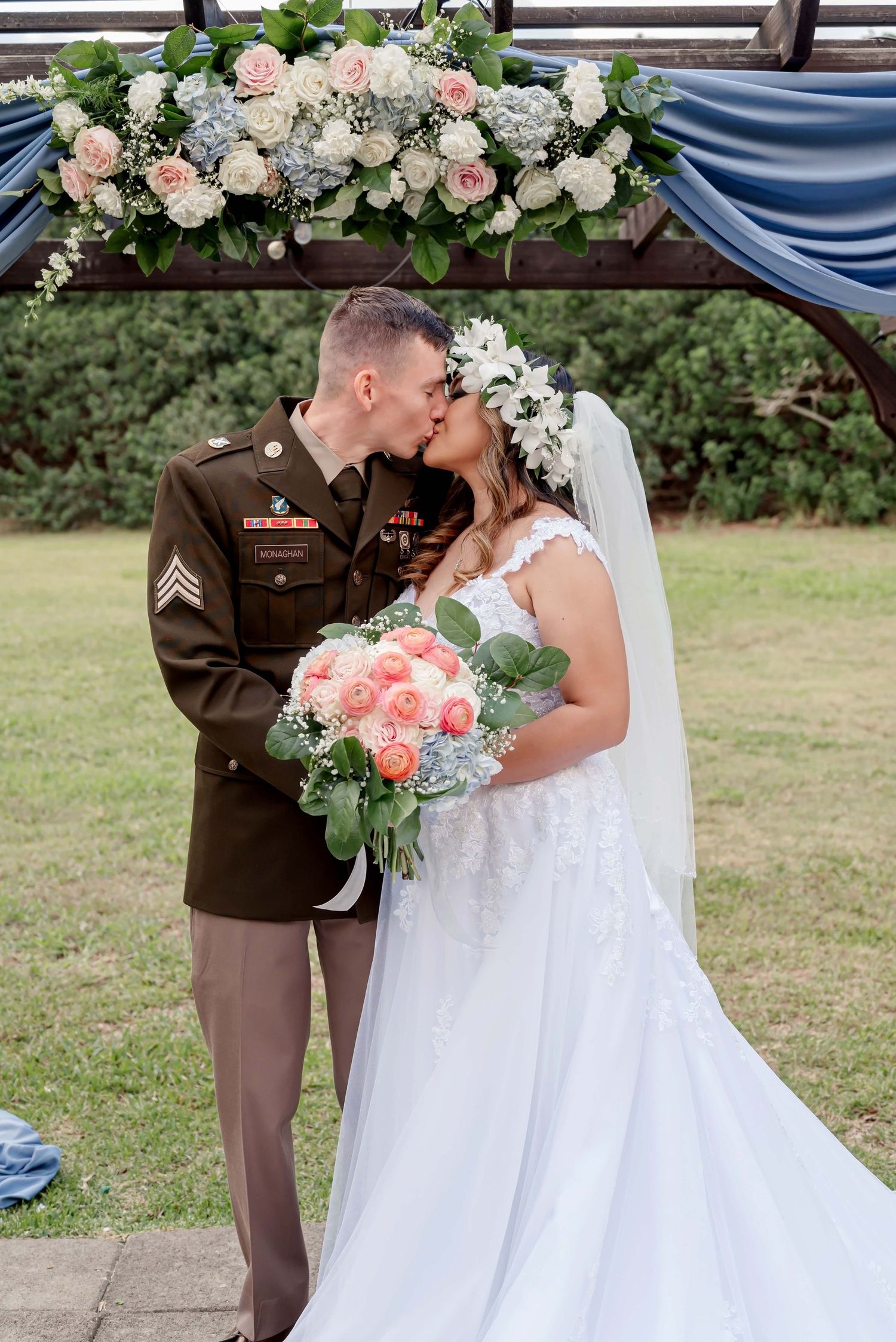 Military couple share an intimate moment during outdoor wedding ceremony with white and pink floral bouquets.