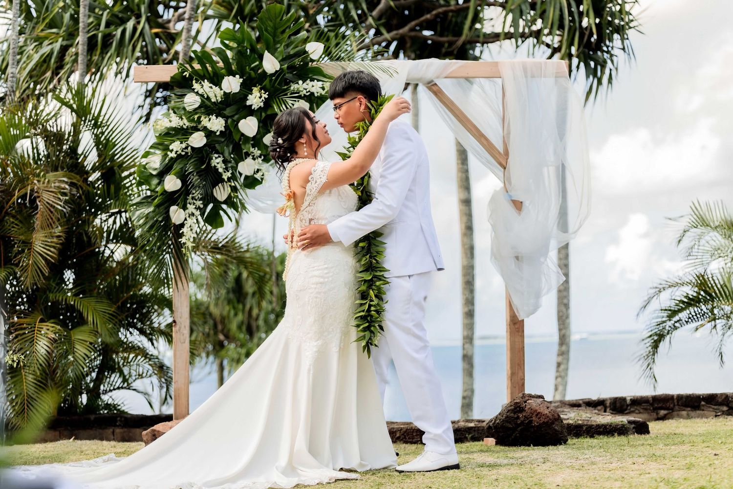 Couple sharing first kiss at tropical outdoor wedding ceremony with white floral arch and palm trees.