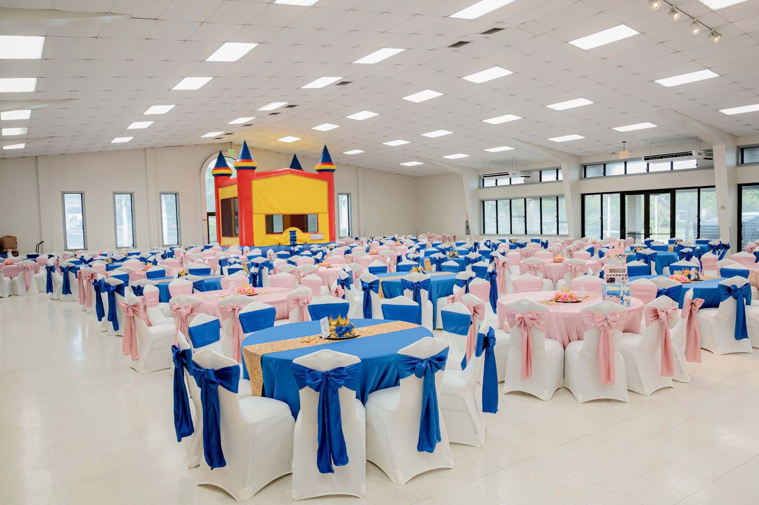 Elegant banquet hall with blue and pink decorated tables, white chair covers, and a colorful bounce castle in the background.