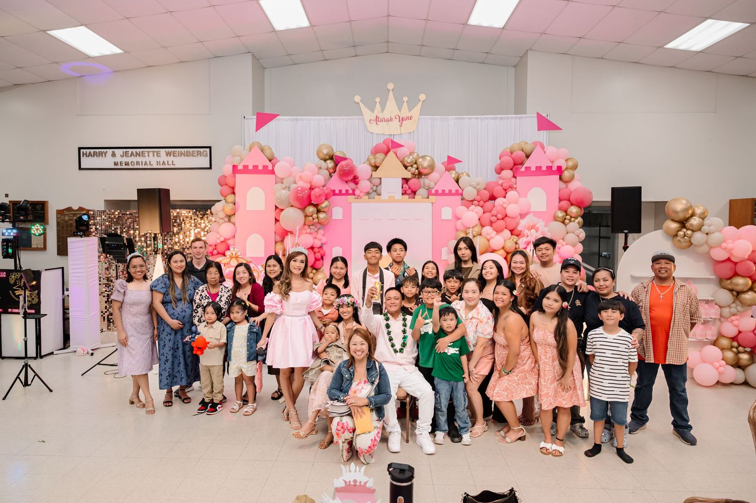 Large group poses at a pink princess-themed birthday party with balloon castle backdrop and gold decorations.