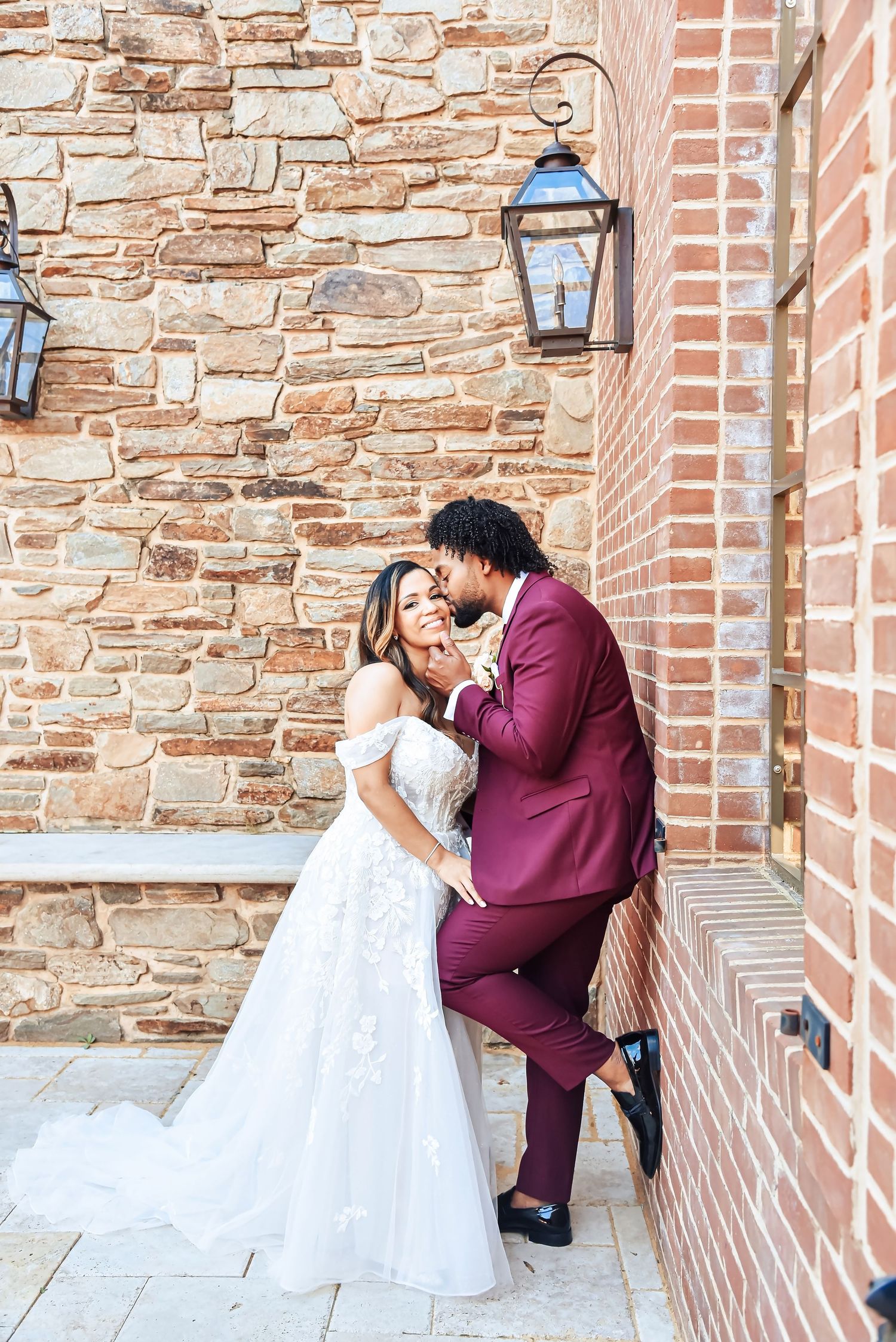 A wedding couple poses romantically against a stone and brick wall with vintage lanterns at a historic venue.