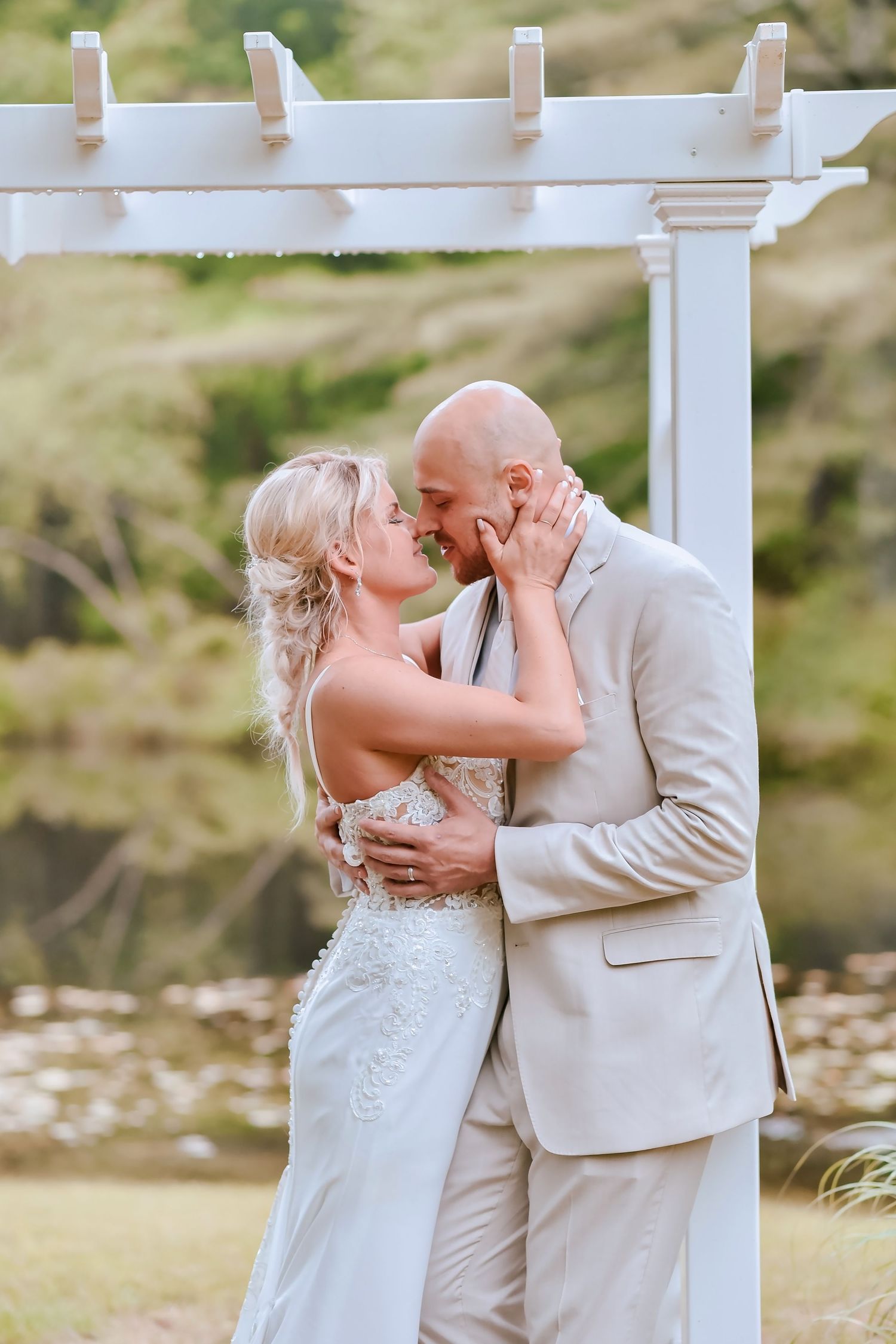 A couple shares a romantic kiss under a white pergola during an outdoor wedding ceremony.