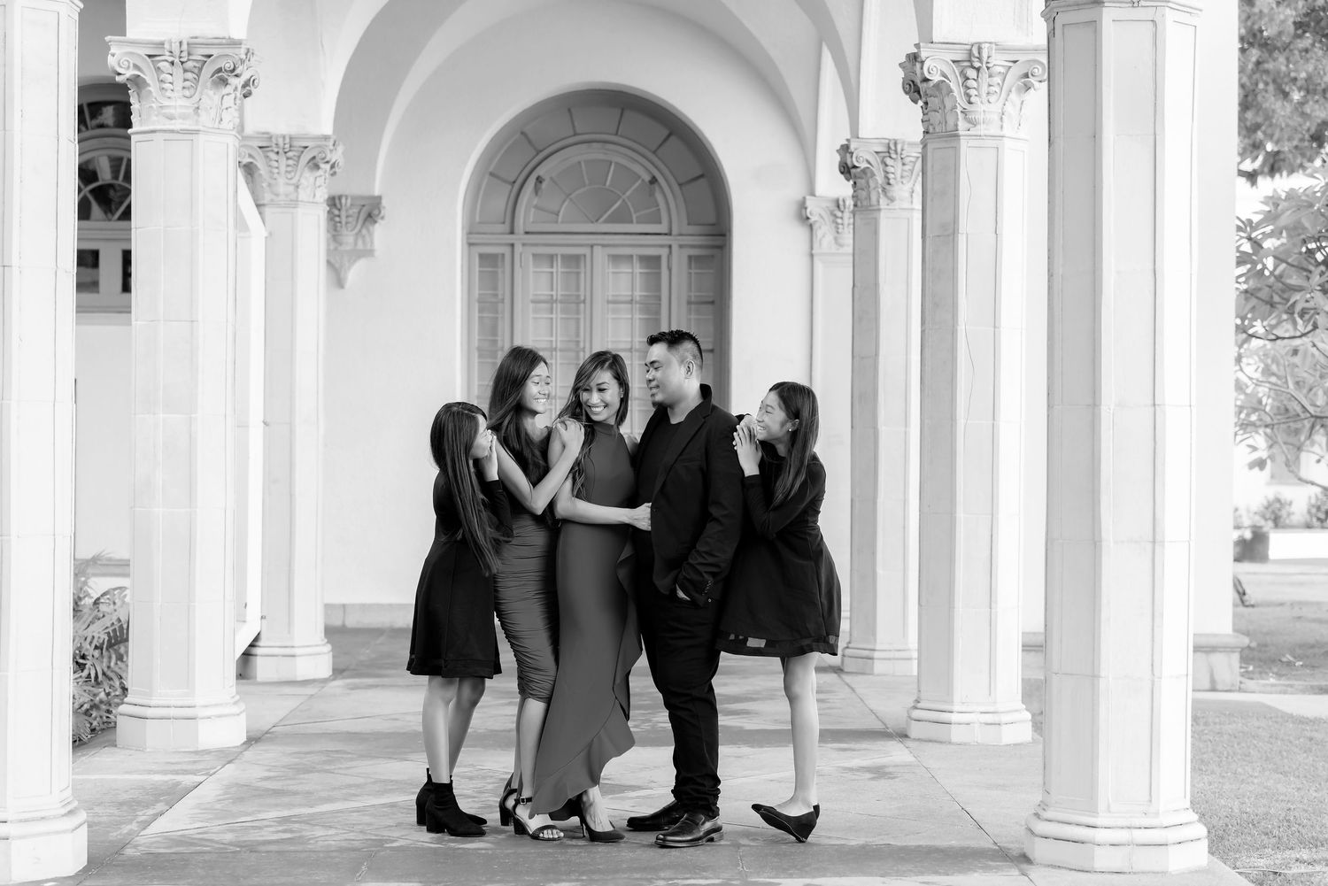 Black and white family portrait of five people embracing and laughing in front of elegant white columns.