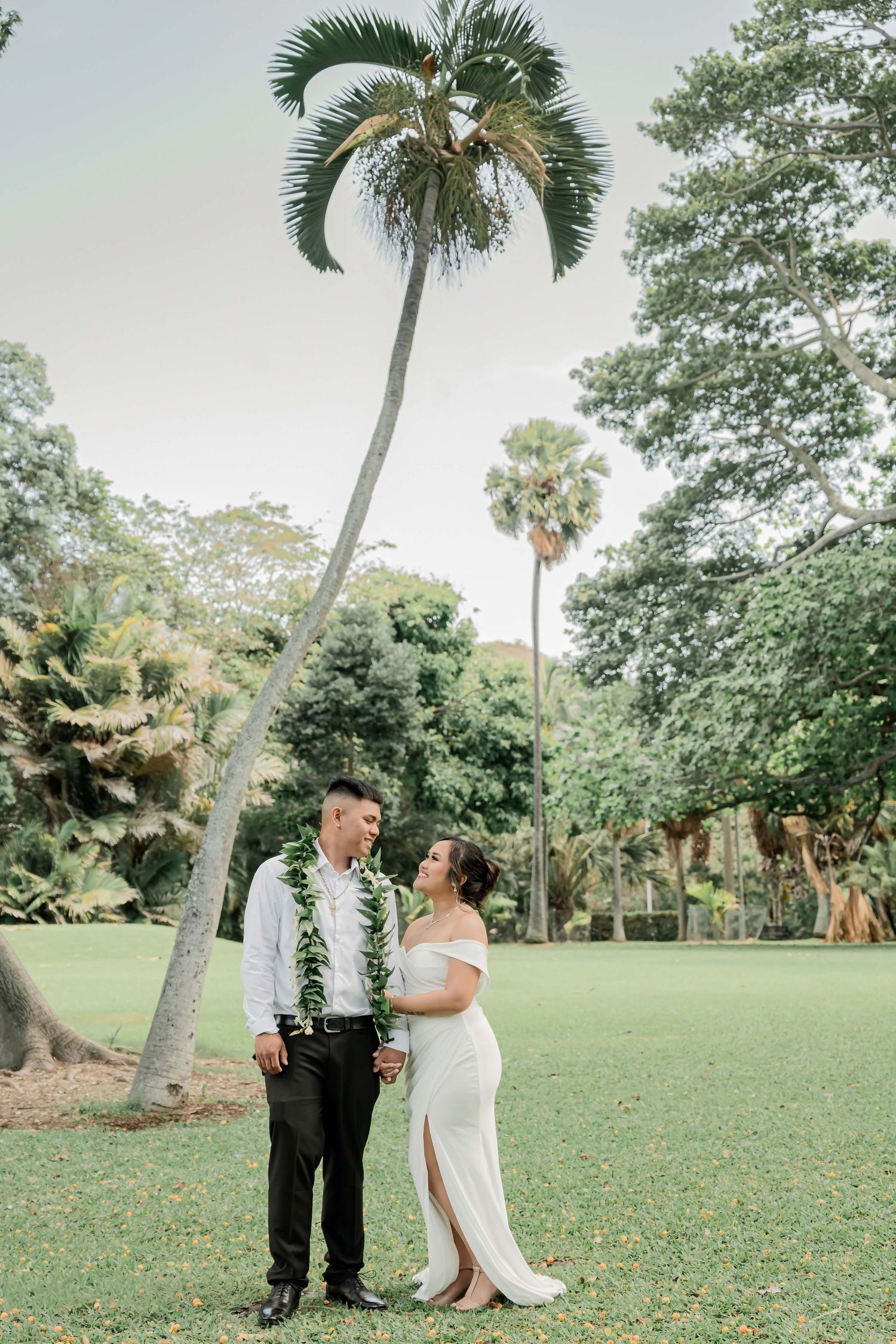 A couple in wedding attire shares a joyful moment together on a lush green lawn surrounded by palm trees and tropical foliage.