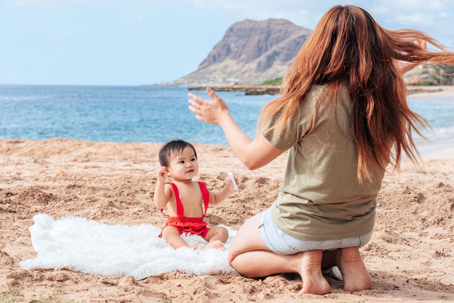 A mother plays with her baby on a sandy beach blanket with blue ocean and mountain views in Hawaii.