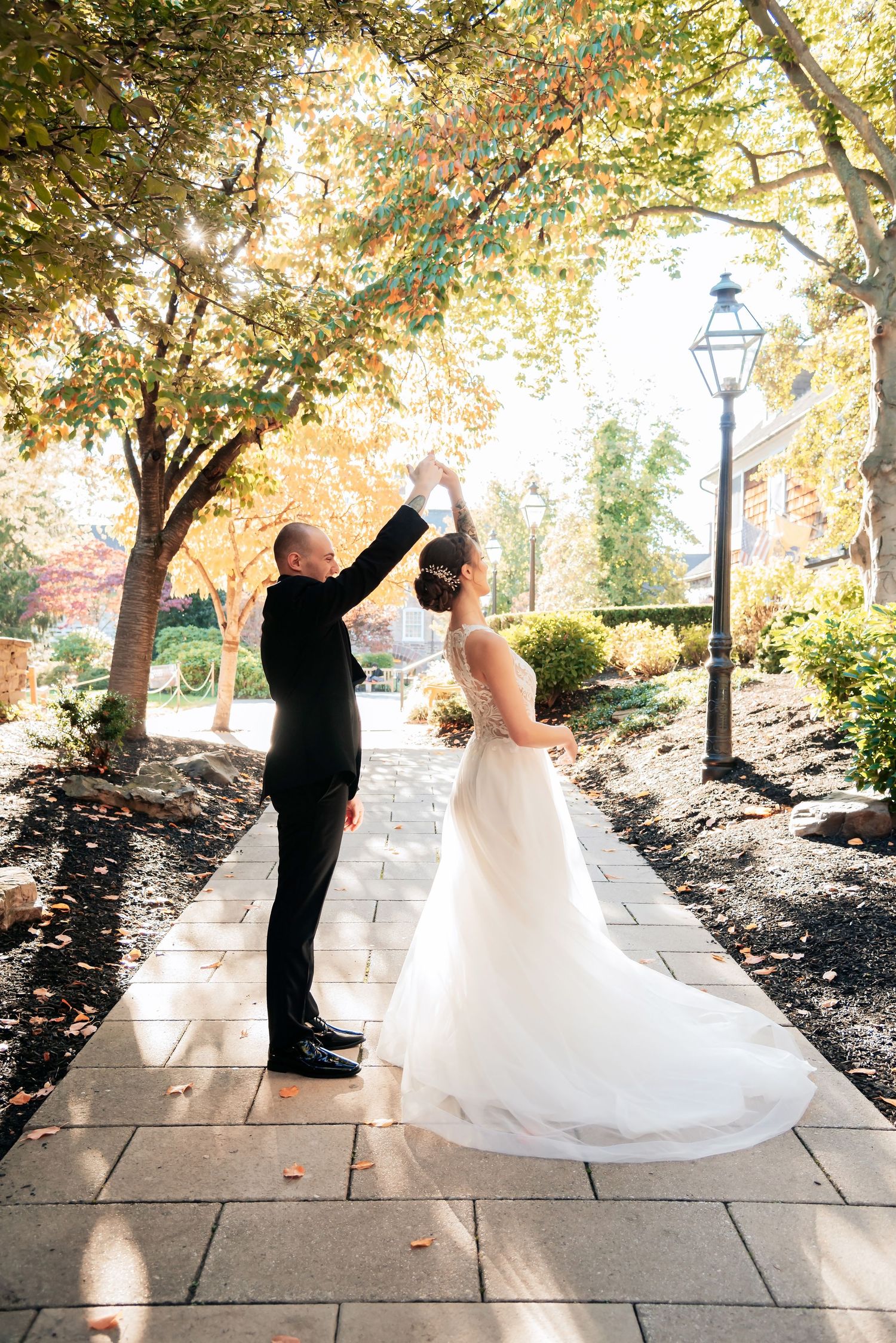 A bride and groom dance together on a stone path surrounded by autumn trees and a vintage lamppost.