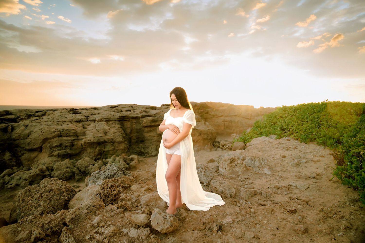 Pregnant woman in white flowing gown poses on rocky cliffside at golden sunset.