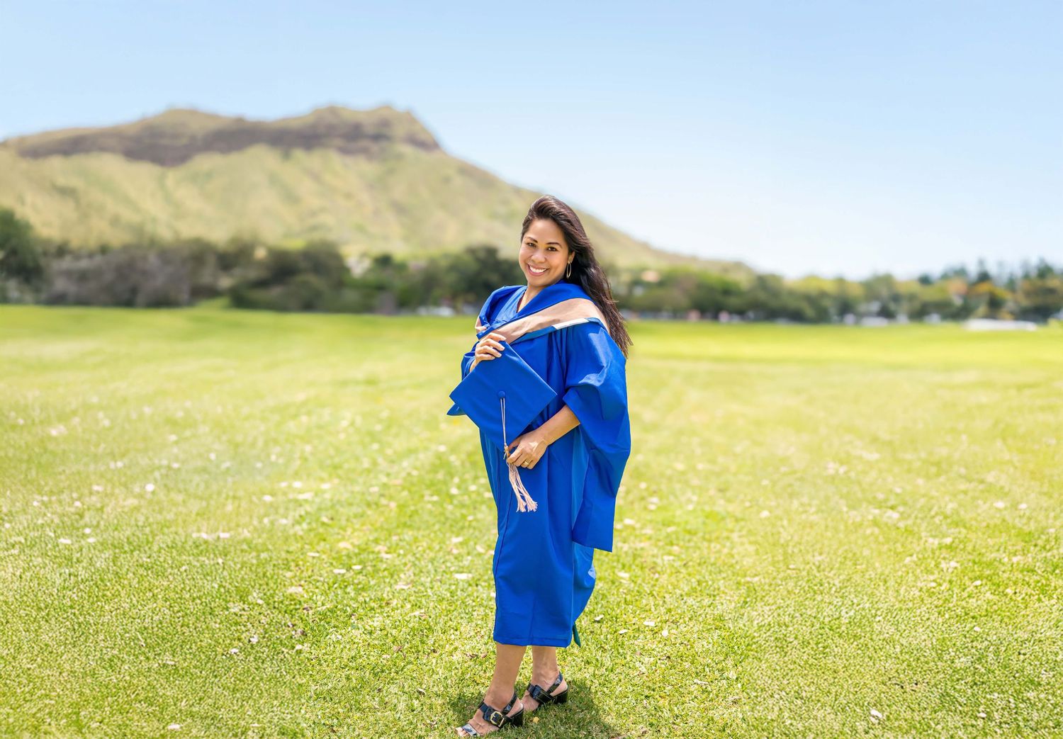 Pregnant woman in blue graduation gown standing in green field with mountain backdrop on sunny day.