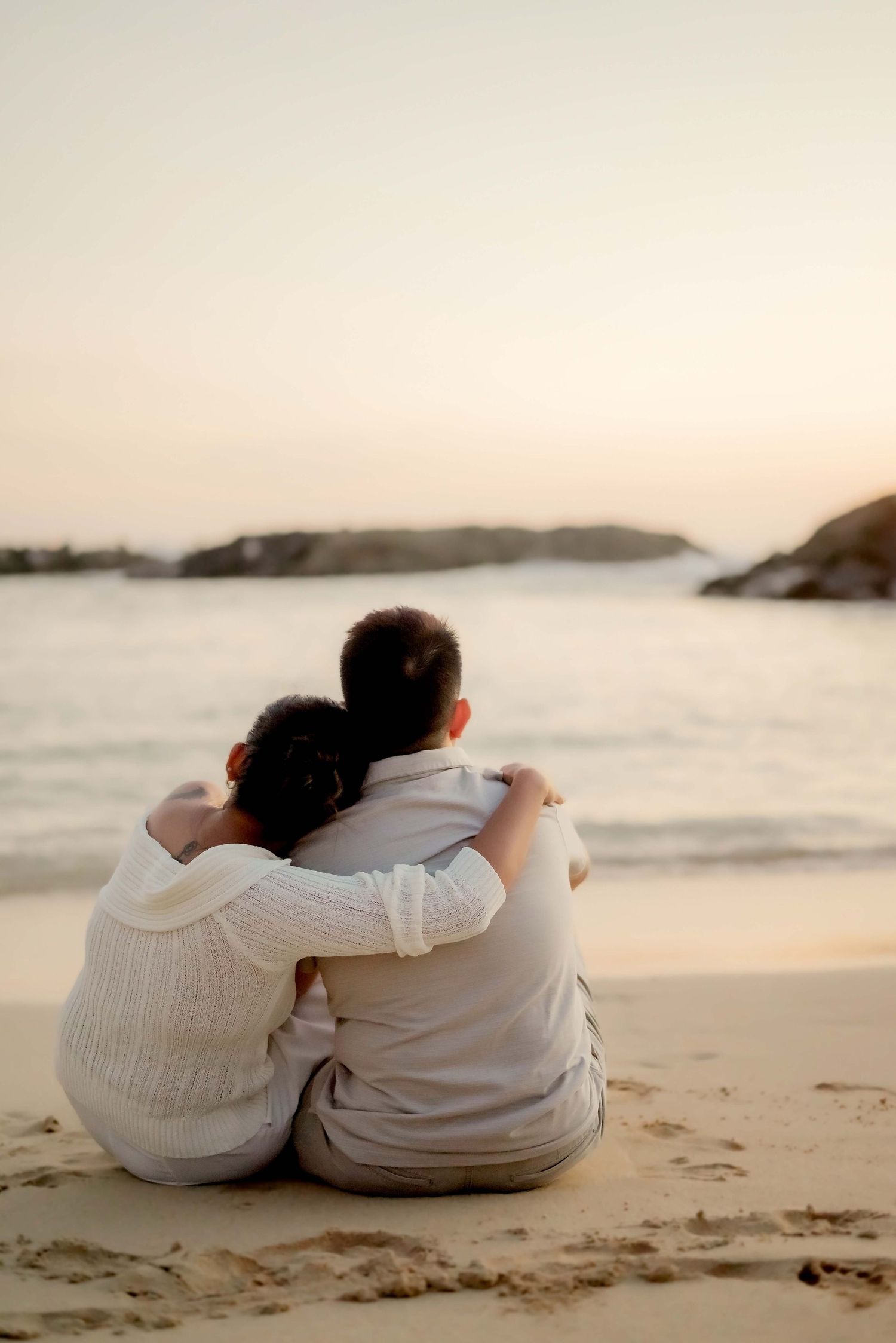Two people in white clothing sit close together on a sandy beach at sunset while watching the ocean waves.