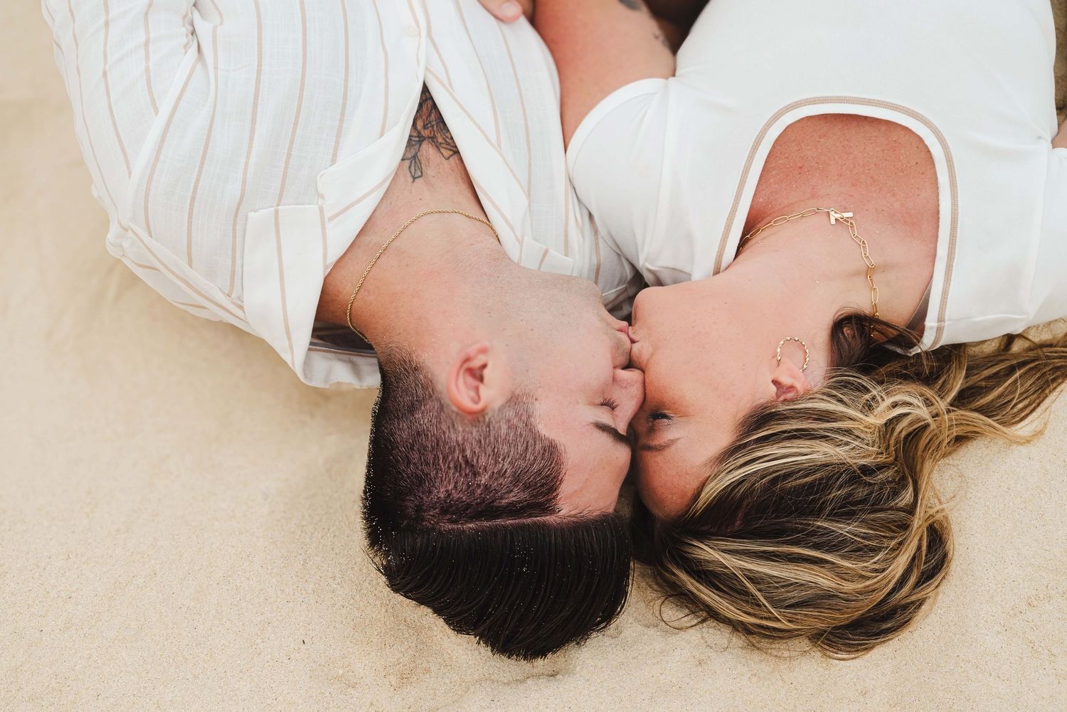 Couple lying on sandy beach sharing a romantic kiss, wearing white outfits in a tender overhead shot.