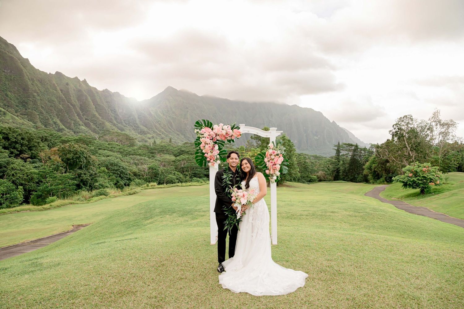 Couple poses under floral arch at Hawaii wedding with lush Ko'olau Mountains backdrop on green lawn.