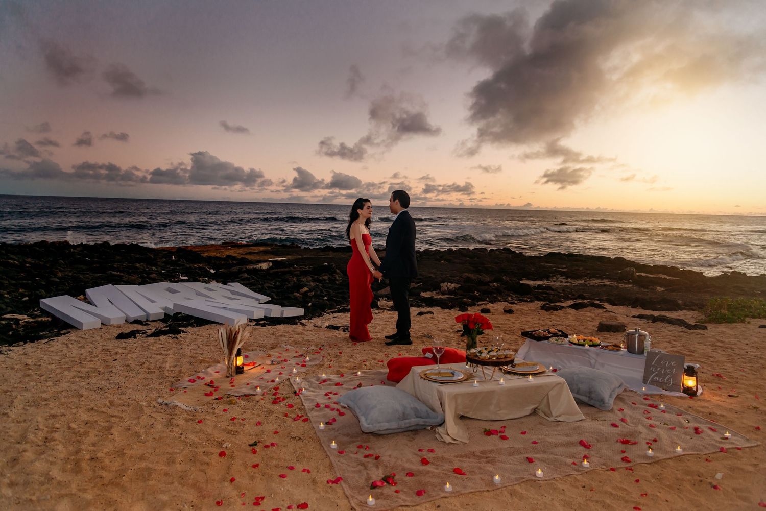 Romantic beach proposal at sunset with rose petals, candles, and cushions by the ocean shoreline.