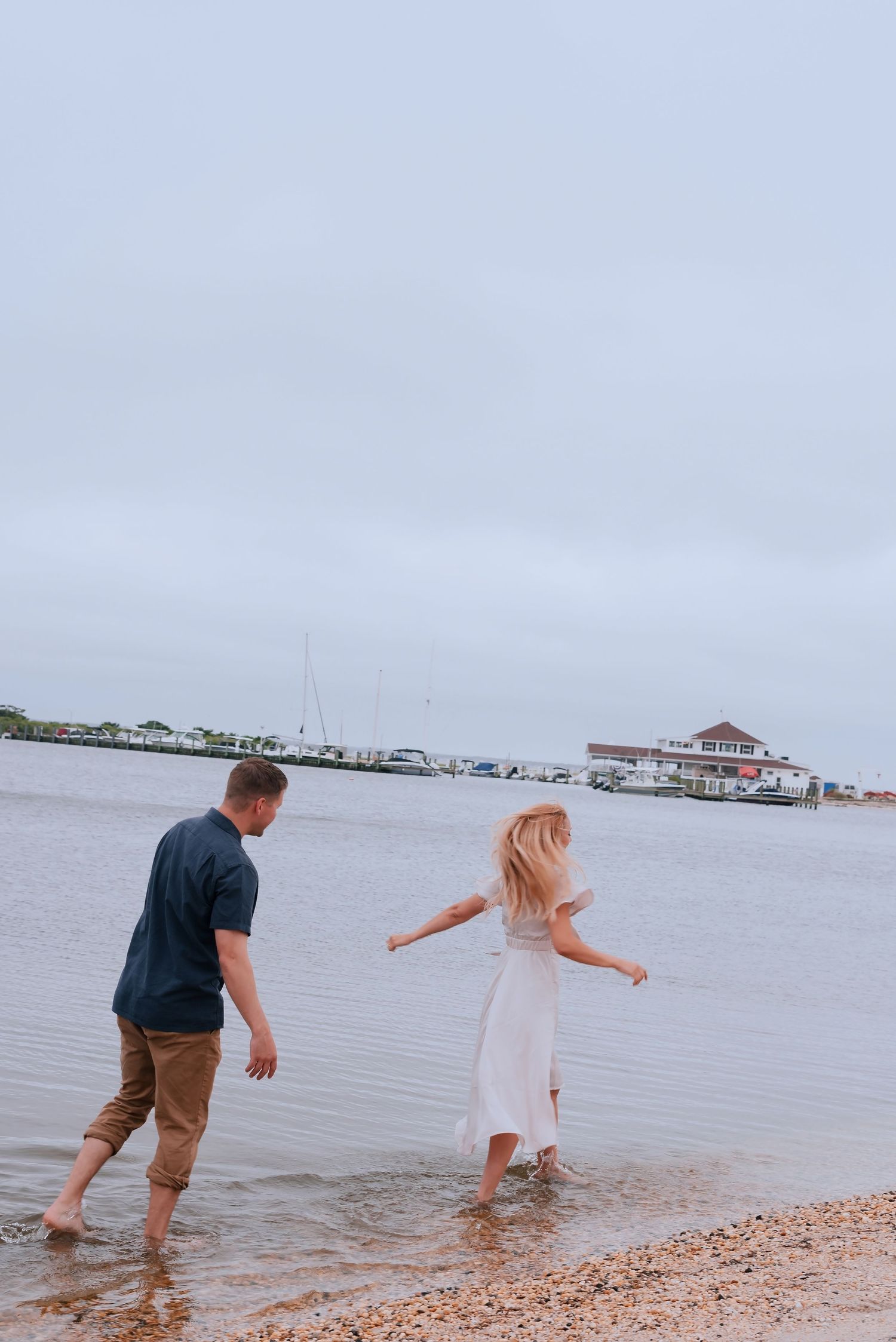 Couple wading barefoot in shallow water at a marina on a cloudy day, woman in white dress, man in navy shirt.