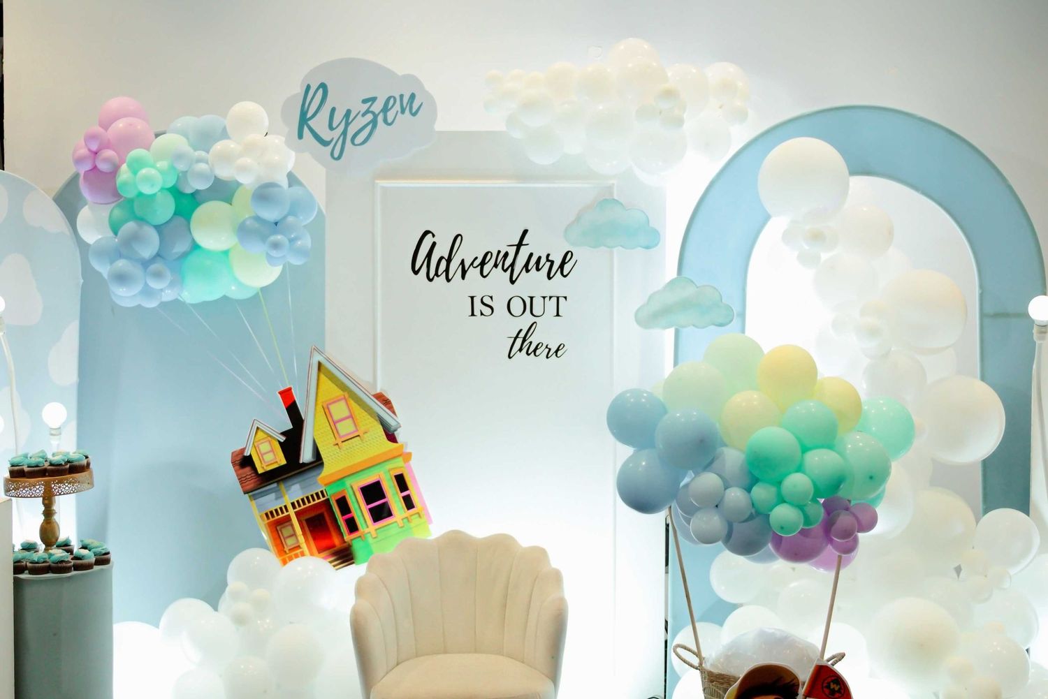 A whimsical baby shower backdrop with pastel balloons, clouds, and a UP-inspired house with the quote Adventure Is Out There.