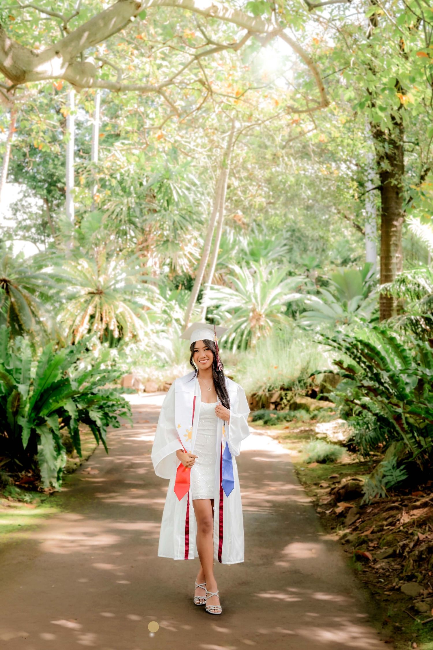 Young woman in white graduation gown and cap walks a lush garden path, smiling, holding colorful honor cords.