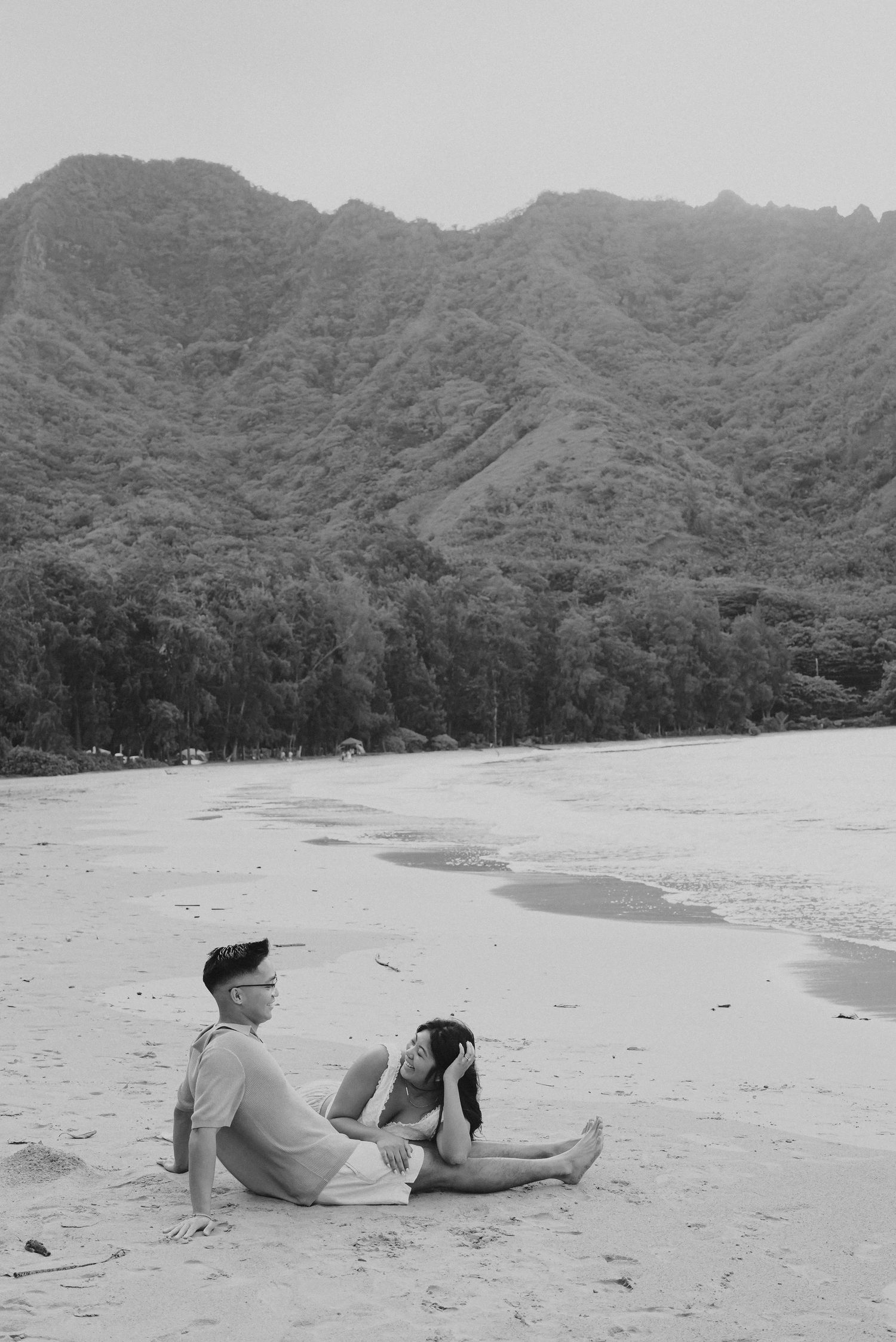 A couple relaxes on a scenic beach with dramatic mountain ridges in the background in black and white.