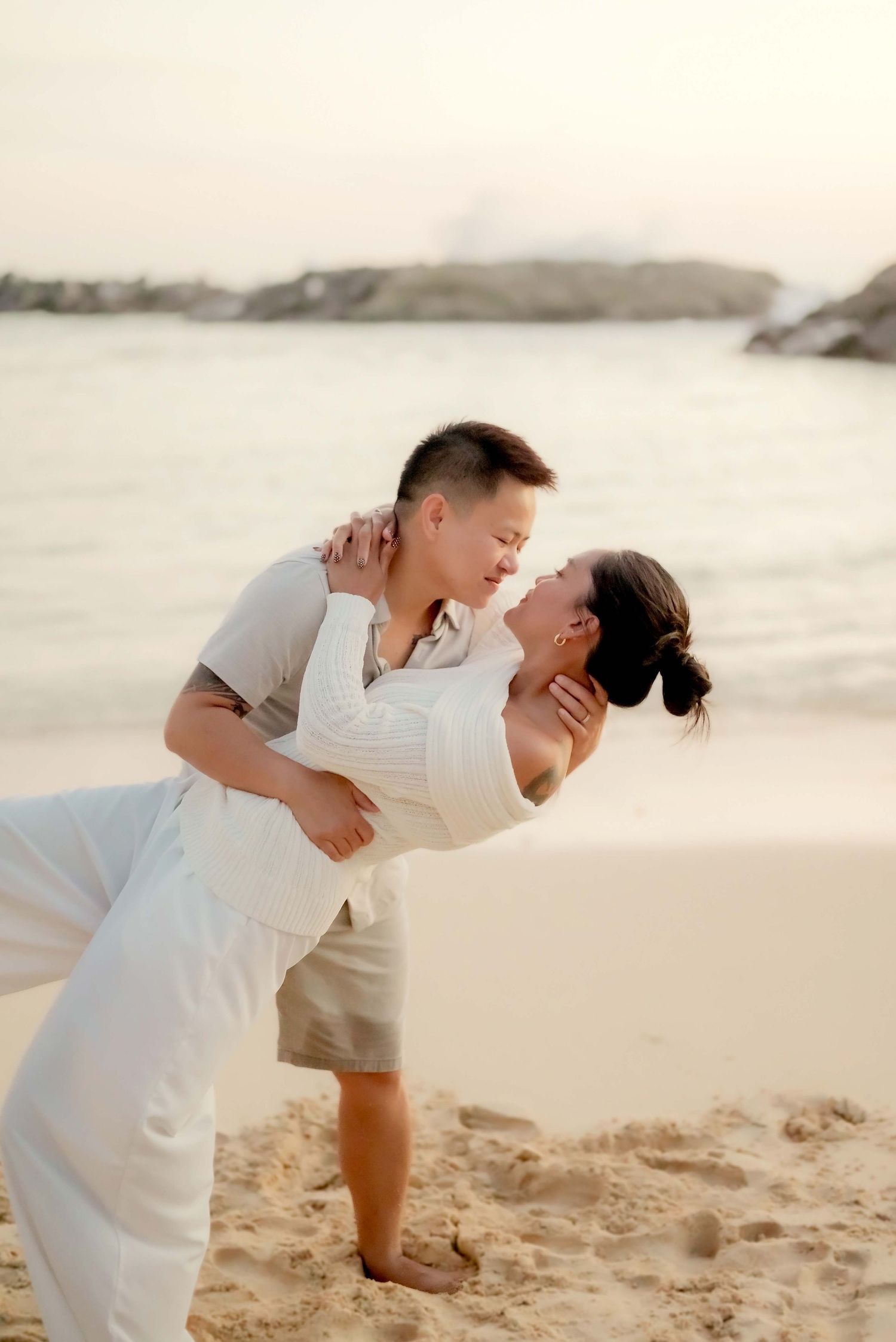 A romantic couple in white outfits share a tender moment while dancing on a sandy beach at sunset.