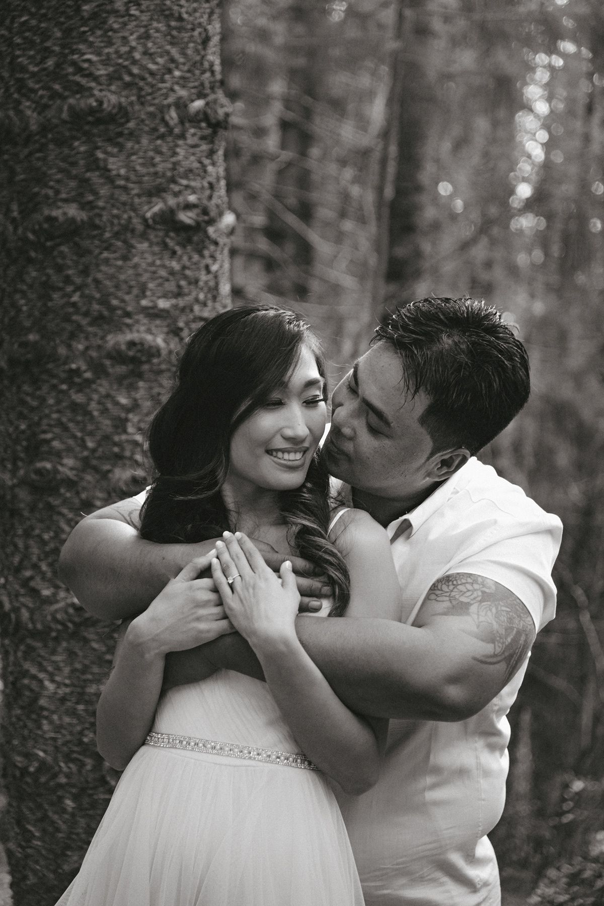 A couple sharing tender romantic moments in a forest setting captured in black and white photography.