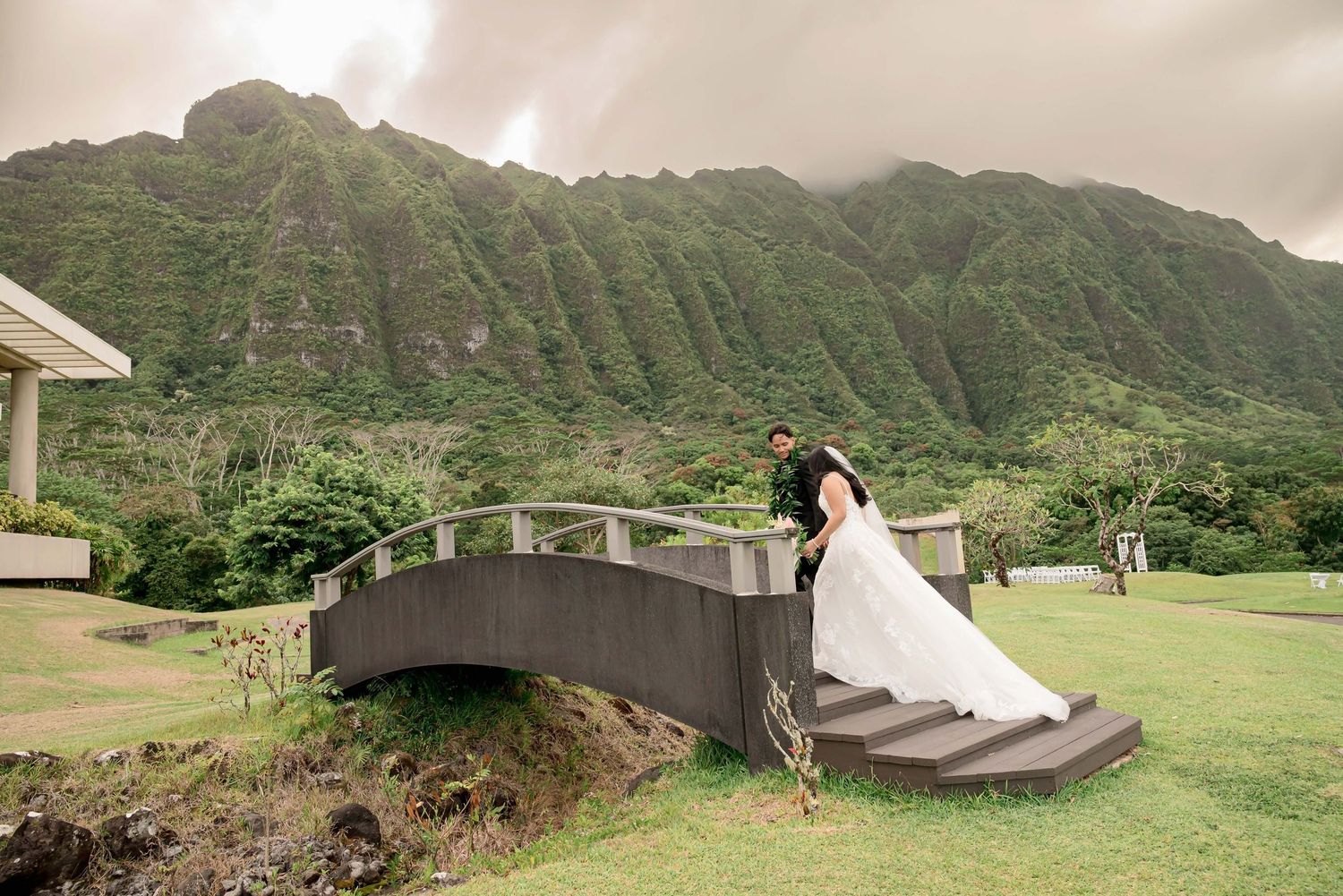 Bride and groom pose on a stone bridge with lush green Ko'olau Mountains backdrop in Hawaii.