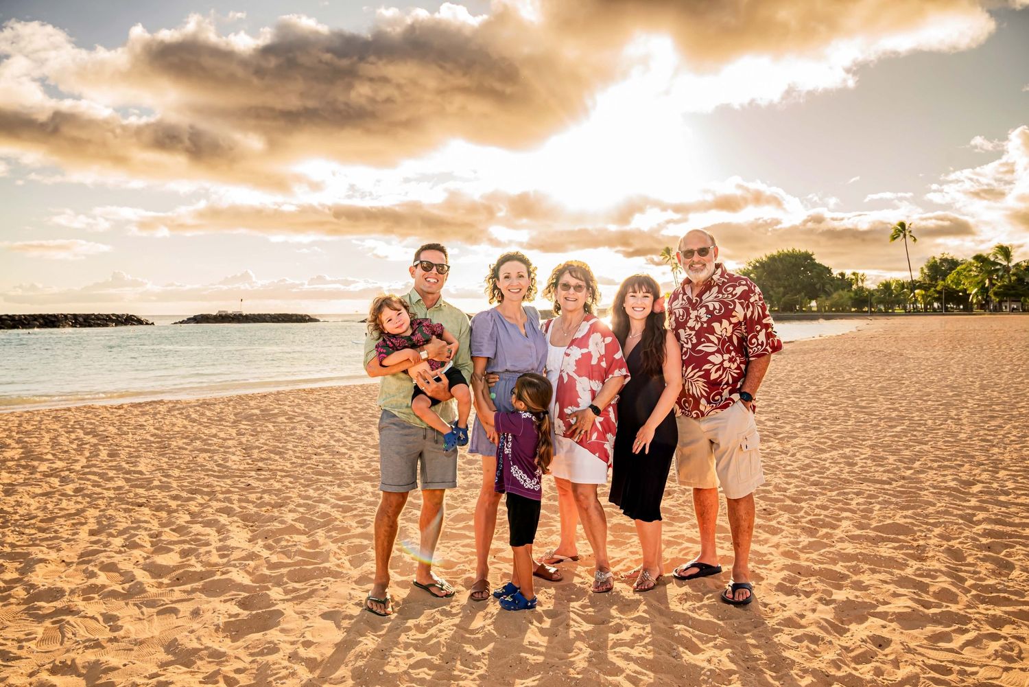 Family of seven posing together on a sandy beach at golden sunset in Hawaii, wearing colorful summer outfits.