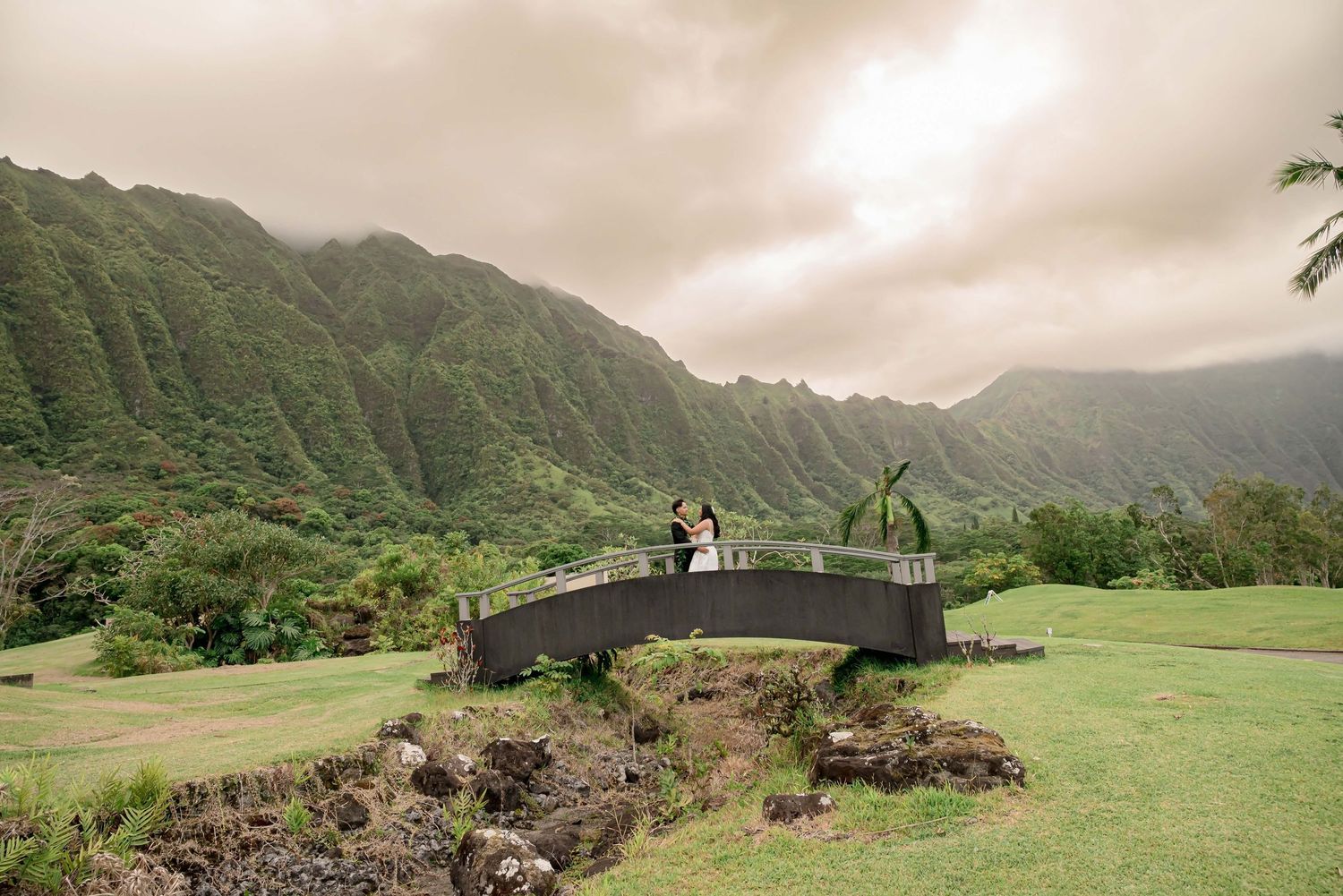 Couple poses on a bridge surrounded by lush green mountains and tropical landscape in Hawaii.