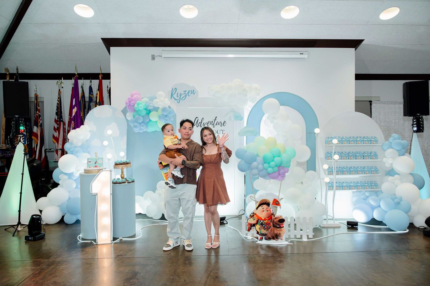 Family posing at a baby's first birthday party with blue balloon arch decorations and festive backdrop.