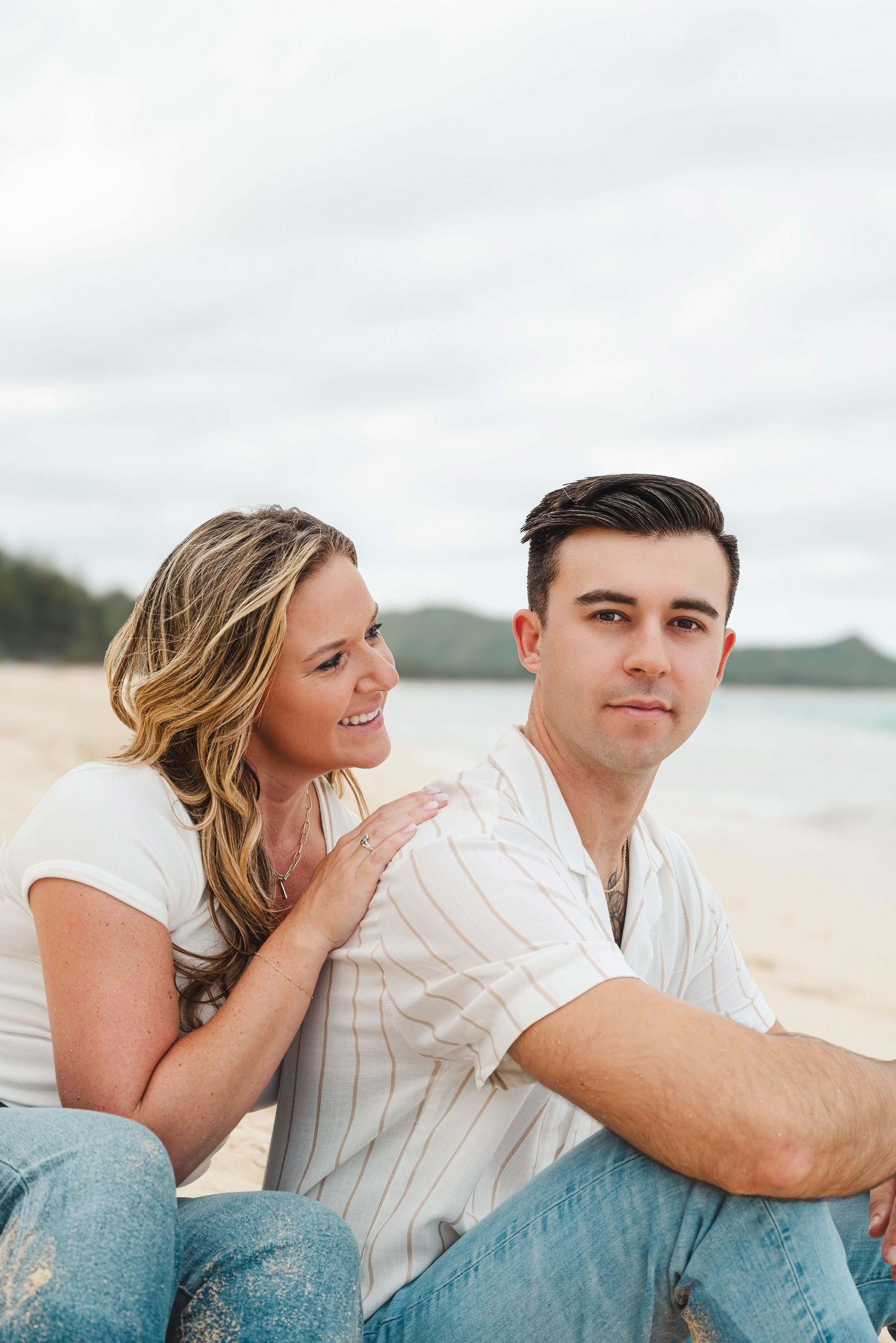 Couple sitting close together on a beach with overcast sky and ocean in the background.