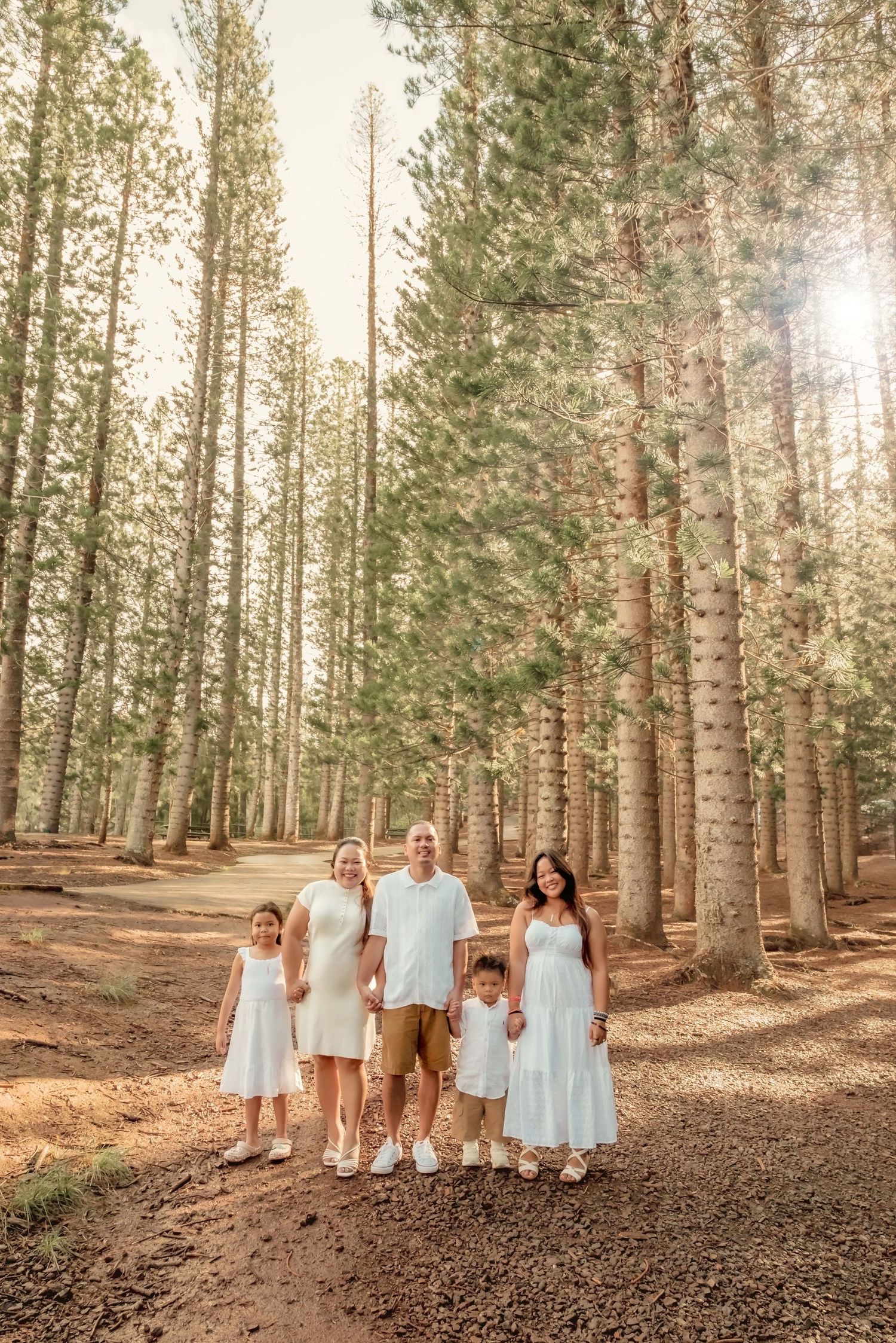 Family of five posing in sunlit pine forest, wearing coordinated white and neutral outfits on a dirt path.