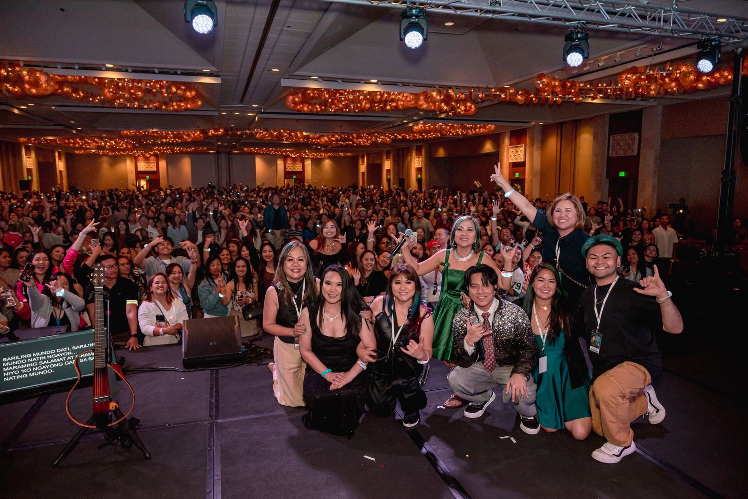 Large crowd at an indoor event venue with group of smiling attendees posing on stage in green and black outfits.