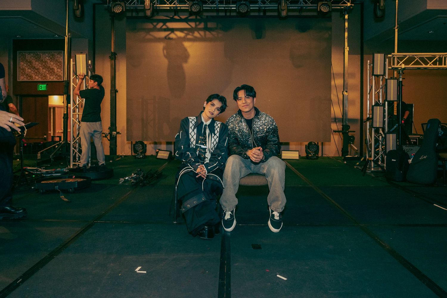 Two young men sitting together in a gym setting, wearing casual streetwear outfits in a moody, dimly lit environment.